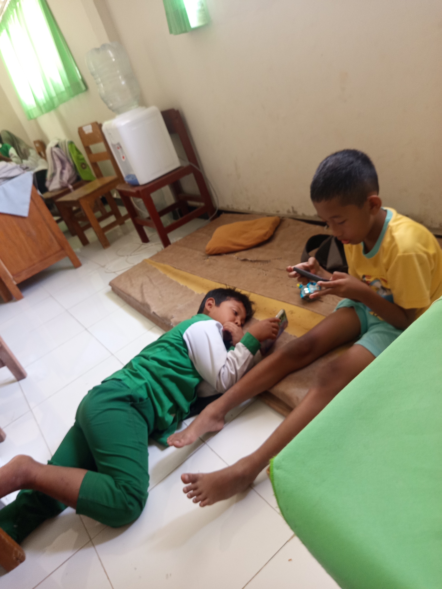 Two young boys are lying on mats on the floor of what appears to be a classroom or common area in Lumutan, Indonesia. The boy on the left is lying on his side, wearing a green and white long-sleeved shirt and green pants, looking at a smartphone. The boy on the right is sitting up, wearing a yellow t-shirt and light blue shorts, also looking at a smartphone. In the background, there are several wooden chairs, a water dispenser with a large bottle on top, and green curtains covering windows. The floor is tiled.