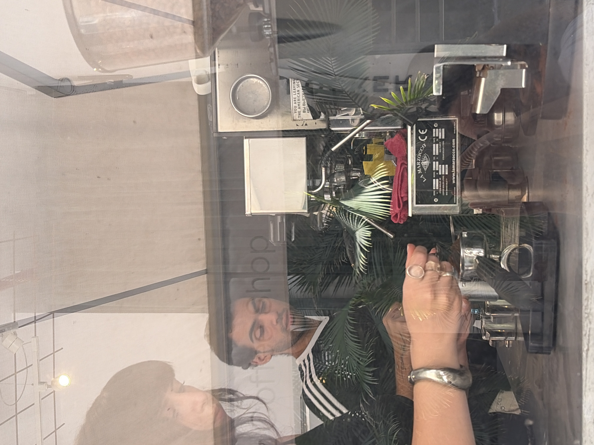 A medium shot captures a reflection in a window, likely a cafe in Tel-Aviv, Israel, showing two individuals, one man and one woman, preparing coffee. The woman on the left, partially visible, wears a large silver bracelet and rings, and is in the process of tamping coffee grounds into an espresso machine. The man, to the right, is wearing a black and white striped Adidas-style shirt, looking down at the machine. In the background, an espresso machine, possibly a La Marzocco, is visible, along with palm fronds and a grinder. The scene is illuminated by overhead lighting. The overall impression is one of a bustling cafe environment.