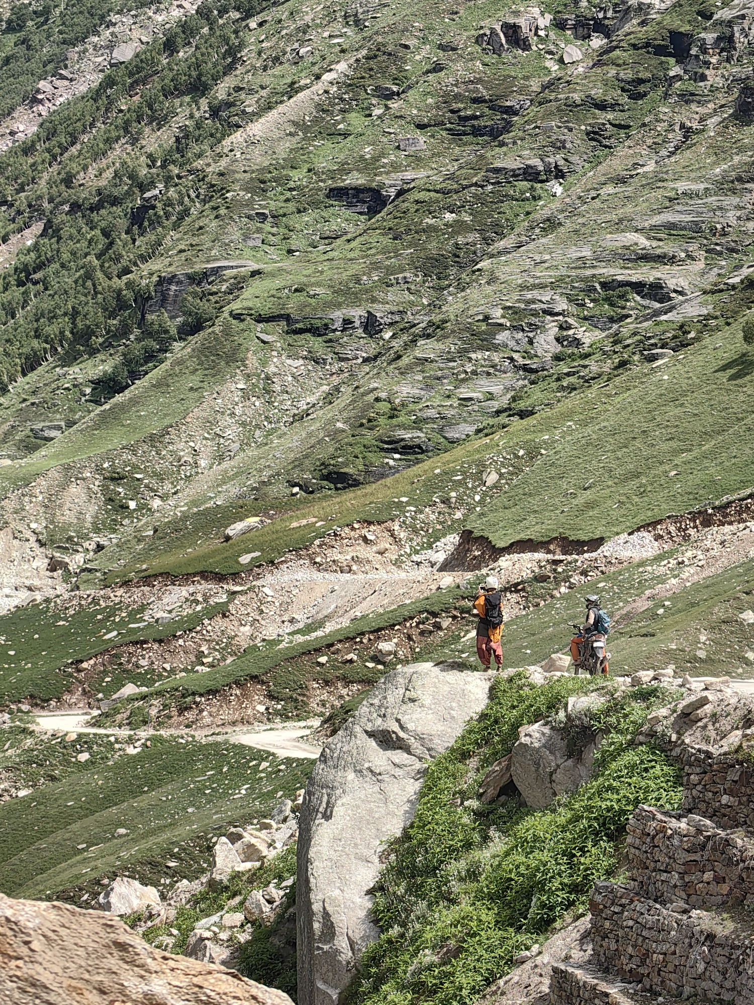 In the mountainous terrain of Burwa, India, two individuals are observed. One person, standing on a rock, wears an orange top and brown pants, with a black backpack. To their right, a second person is seated on an orange motorcycle, also equipped with a backpack. The environment is characterized by steep, grassy slopes interspersed with rocky outcrops and a dirt road winding through the landscape. The vegetation is predominantly grass and some scattered trees on the upper slopes. The scene appears to be a remote, natural setting within the Indian Himalayas.