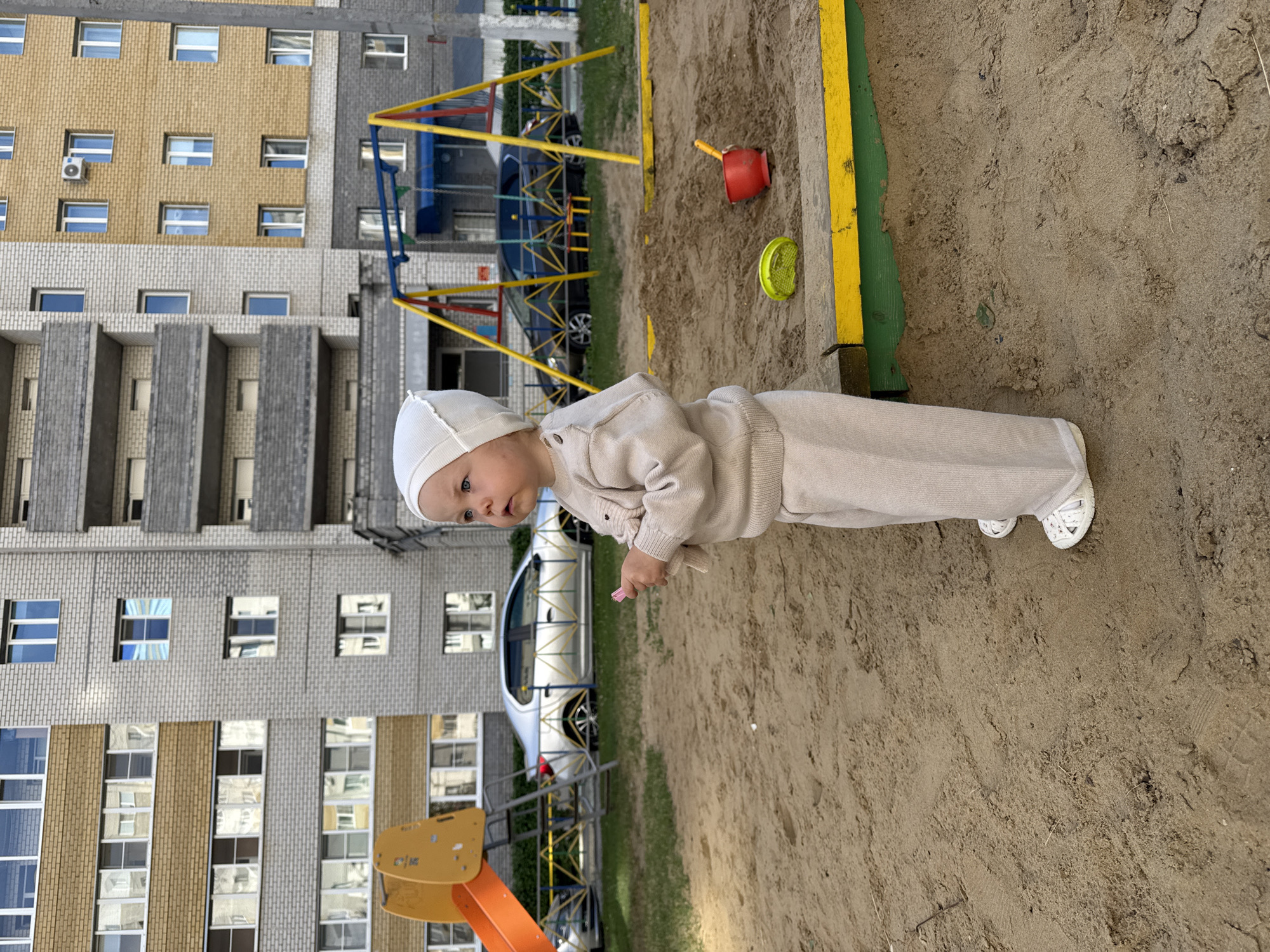 This is a full shot, taken outdoors, of a young child standing in a sandy area. The child is wearing a light-colored outfit consisting of a hat, a long-sleeved shirt, and pants. White shoes are visible on their feet. The child's gaze is directed slightly to the left, and their right hand is extended forward, holding a small pink object. In the background, there's a playground with a yellow and green border around the sandpit, and further back, a multi-story residential building with many windows. A dark car is parked next to the playground equipment. The sky is not visible, but the lighting suggests it's daytime. The scene appears to be a residential area, possibly a playground in a courtyard. The image is rotated 90 degrees counterclockwise.