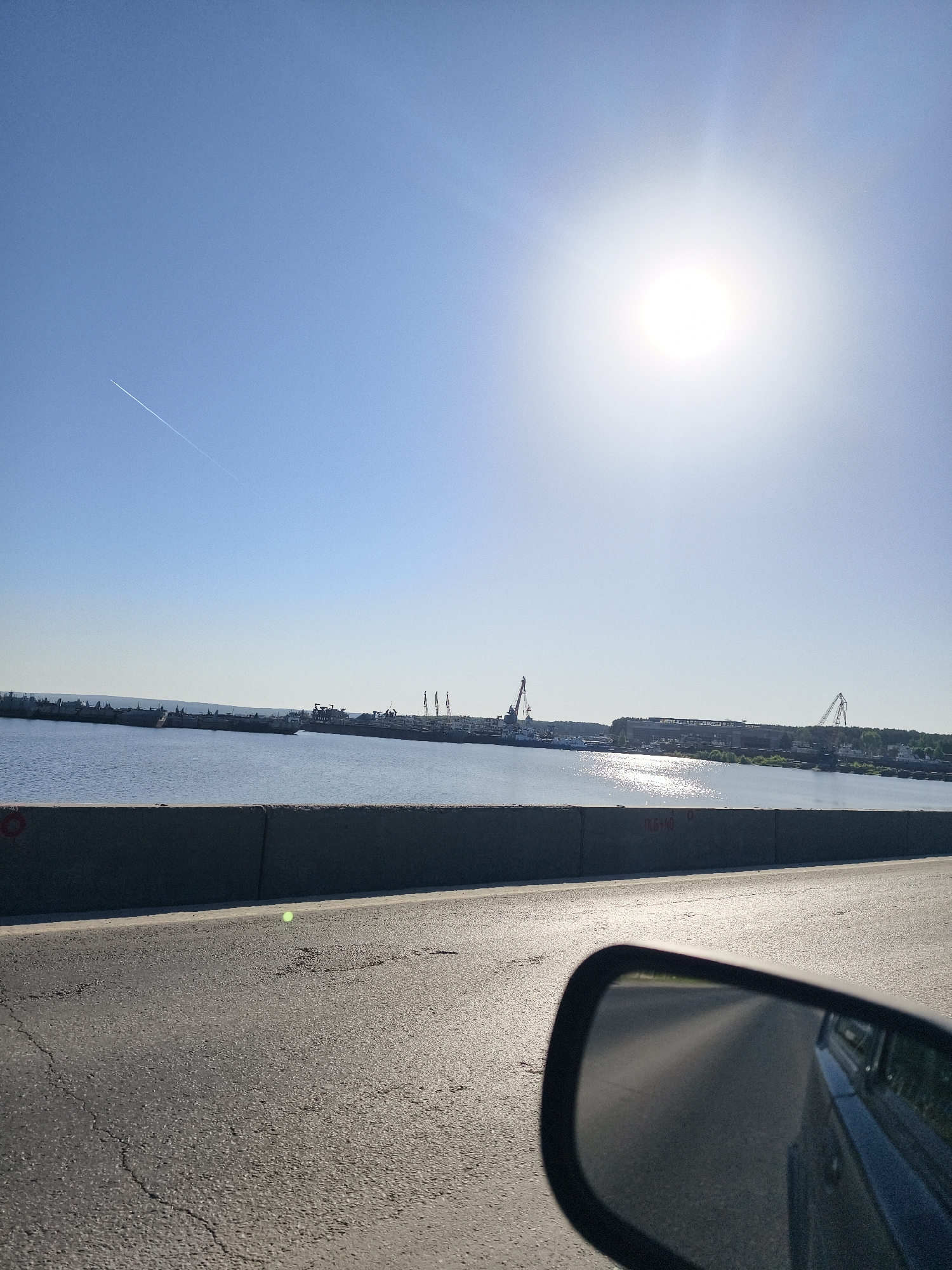 The image presents a daytime view from a road, likely a bridge, in Chaikovsky, Russia. The sky is bright and clear with the sun positioned high, creating glare. A contrail is visible in the upper left portion of the sky. In the distance, a body of water is bordered by a shoreline with industrial facilities, including cranes. A concrete barrier runs along the edge of the road, and a car's side mirror is visible in the foreground, reflecting part of the car. The road surface is asphalt.