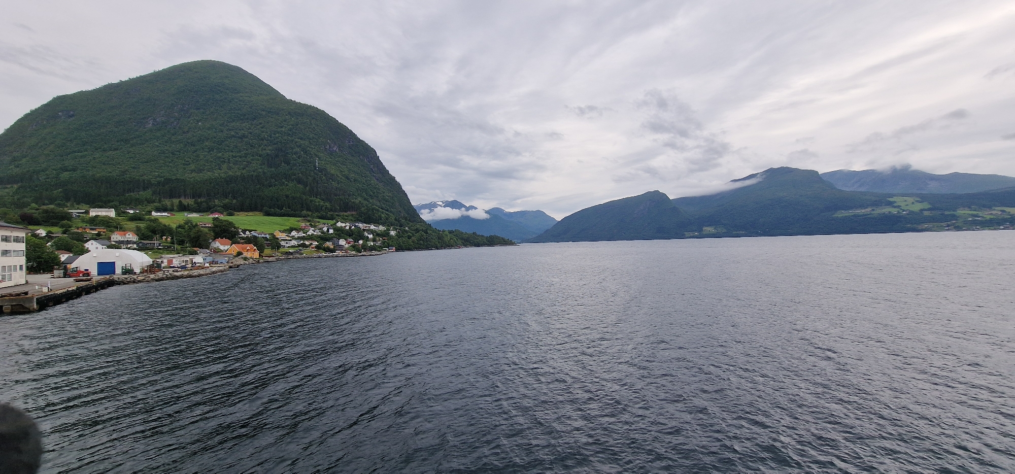 This media file captures a serene view of a fjord in Volda, Norway. The scene is dominated by a calm, dark body of water in the foreground, reflecting the overcast sky. In the mid-ground, a charming coastal village nestles at the foot of a verdant, tree-covered mountain. The village features a cluster of houses with colorful roofs, hinting at a residential area. Further along the coastline, industrial buildings with white and blue accents, along with some docks, suggest a small port or industrial zone. Red machinery and some parked vehicles are visible near the buildings.
The background is characterized by a series of majestic, mist-shrouded mountains, their slopes also covered in dense green forests. The sky is heavily clouded, with streaks of lighter clouds suggesting movement and a potentially cool, damp atmosphere. The overall mood is one of peaceful solitude and natural beauty. The time of day appears to be daytime, though the overcast conditions create a diffused light. The location is clearly identified as Volda, Norway, and the landscape features are typical of a Norwegian fjord. No people are explicitly visible, and no text is present in the image.