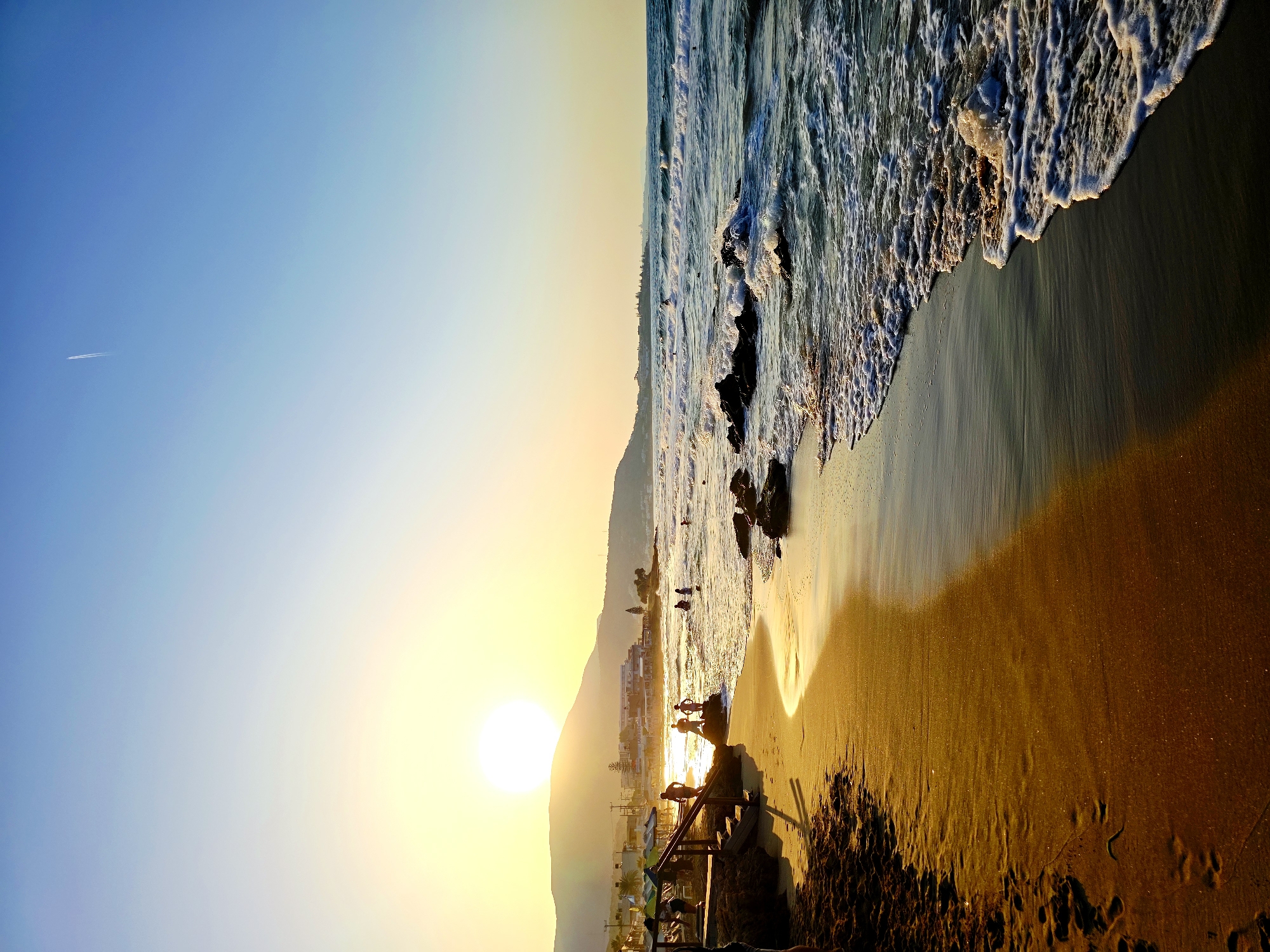This is a landscape photograph of a beach in Malia, Greece during sunset. The sun, a large, bright orb, is low on the horizon behind a silhouette of hills and buildings. The sky is a gradient of pale yellow near the sun, transitioning to a clear blue higher up, with a faint airplane contrail visible. The beach itself is golden and wet near the waterline, reflecting the warm light of the setting sun. Footprints are visible in the dry sand on the left side of the image. Waves are breaking on the shore, creating white foam that washes over dark rocks scattered near the waterline. Several people are present on the beach and in the shallow water. Some are standing on the sand, while others are wading or swimming in the sea. A wooden staircase with umbrellas is situated on the left side of the beach, suggesting it's part of a beach resort or public access point. The overall scene depicts a serene and picturesque sunset on a Greek beach.