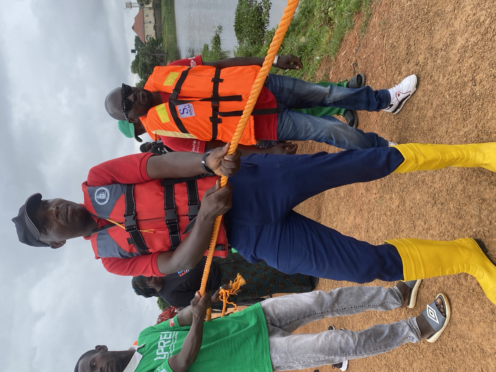 In Kawo, Nigeria, under an overcast sky, a group of individuals are engaged in a tug-of-war with a thick, orange rope near a body of water. Two men in the foreground are pulling the rope with significant effort, their bodies taut. The man on the left wears a green t-shirt with "EPREI" printed on it, grey jeans, and grey slides. He has a watch on his left wrist. The man on the right is wearing a red shirt, blue pants, and bright yellow rubber boots. He also wears a red life vest with grey accents and black buckles. Behind them, another man in an orange life vest with "SOLAS" printed on it and sunglasses stands with his hands on the rope. His attire includes a red t-shirt and blue jeans. Further in the background, other individuals are visible, including a woman wearing a patterned headscarf and dress, and a man in a green cap. The ground is a reddish-brown dirt path, and the water's edge is lined with green vegetation. In the distance, buildings with red roofs and a water tower can be seen.