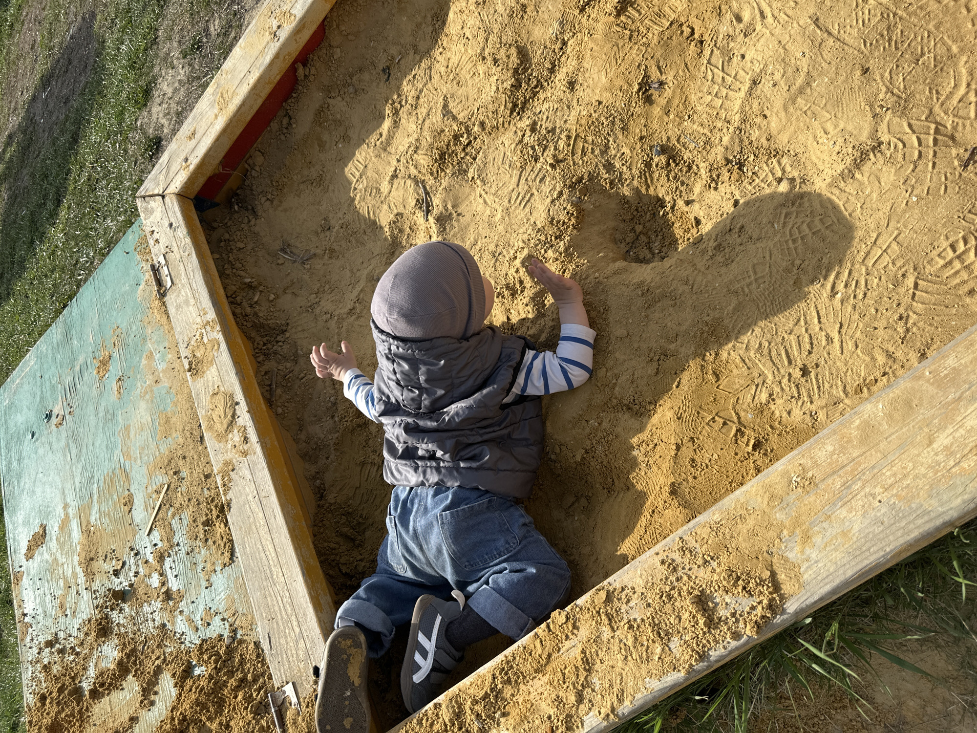 The image shows a child lying in a sandbox. The sandbox is constructed of wooden planks, with one side painted a pale green. The child is wearing a gray hooded hat, a gray vest over a striped long-sleeved shirt, and rolled-up blue jeans. The child's hands are outstretched, pressing into the sand. The sand is marked with footprints and other impressions. The sandbox is situated outdoors, as grass is visible around its perimeter. The city and state are unknown.