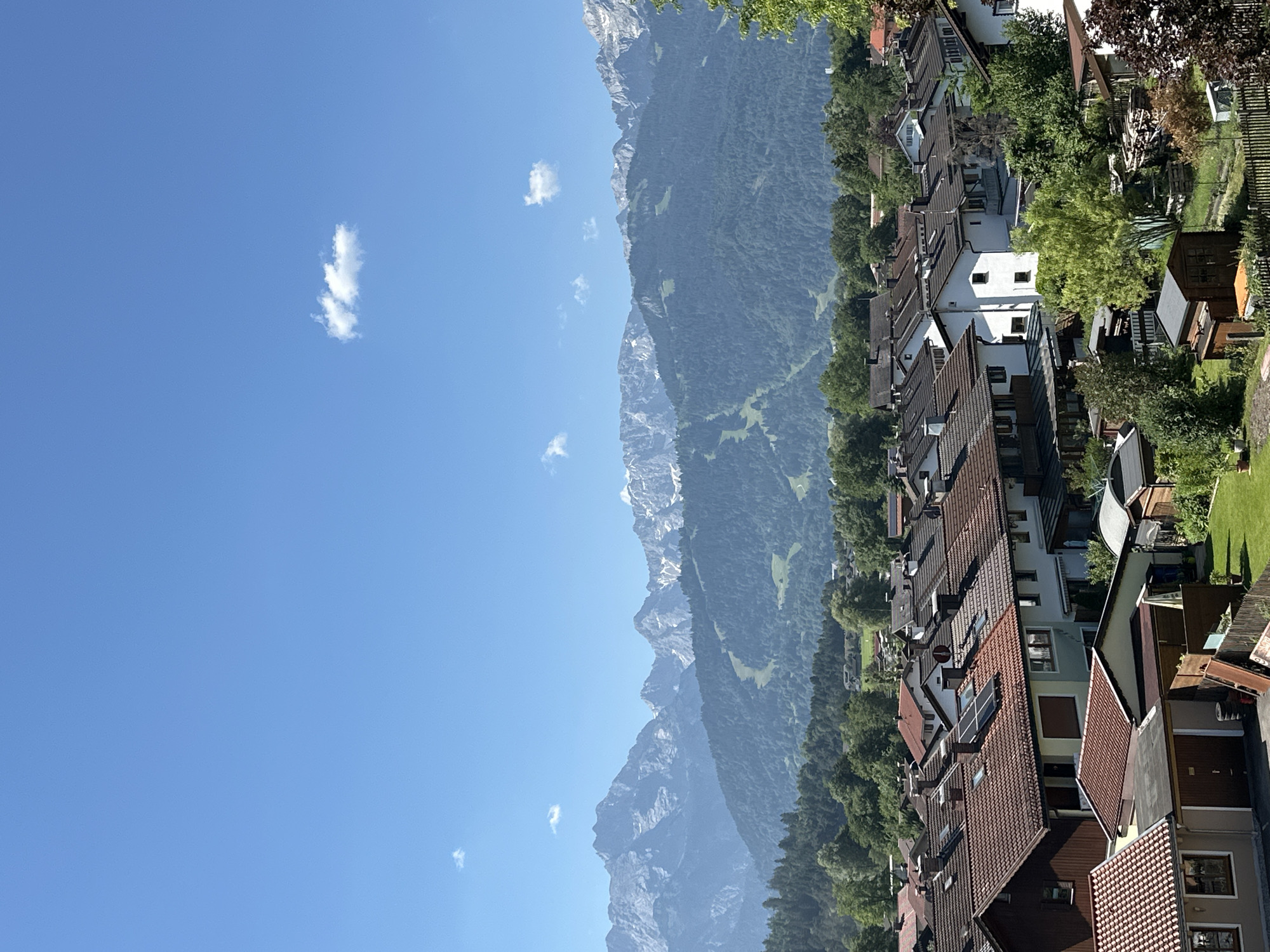 The image shows a residential area in Garmisch-Partenkirchen, Germany, with a backdrop of the Alps. In the foreground, a collection of houses with red-tiled roofs and white walls are visible. Some houses have solar panels installed on their roofs. Numerous trees and green spaces are interspersed among the buildings, suggesting a suburban or semi-rural setting. The middle ground features a dense forest covering the lower slopes of the mountains. The background is dominated by the majestic Bavarian Alps, with their snow-capped peaks piercing a clear blue sky. A single, small white cloud floats in the sky. The scene depicts a sunny day with excellent visibility, offering a panoramic view of the town and its natural surroundings. No people or animals are visible in the image.