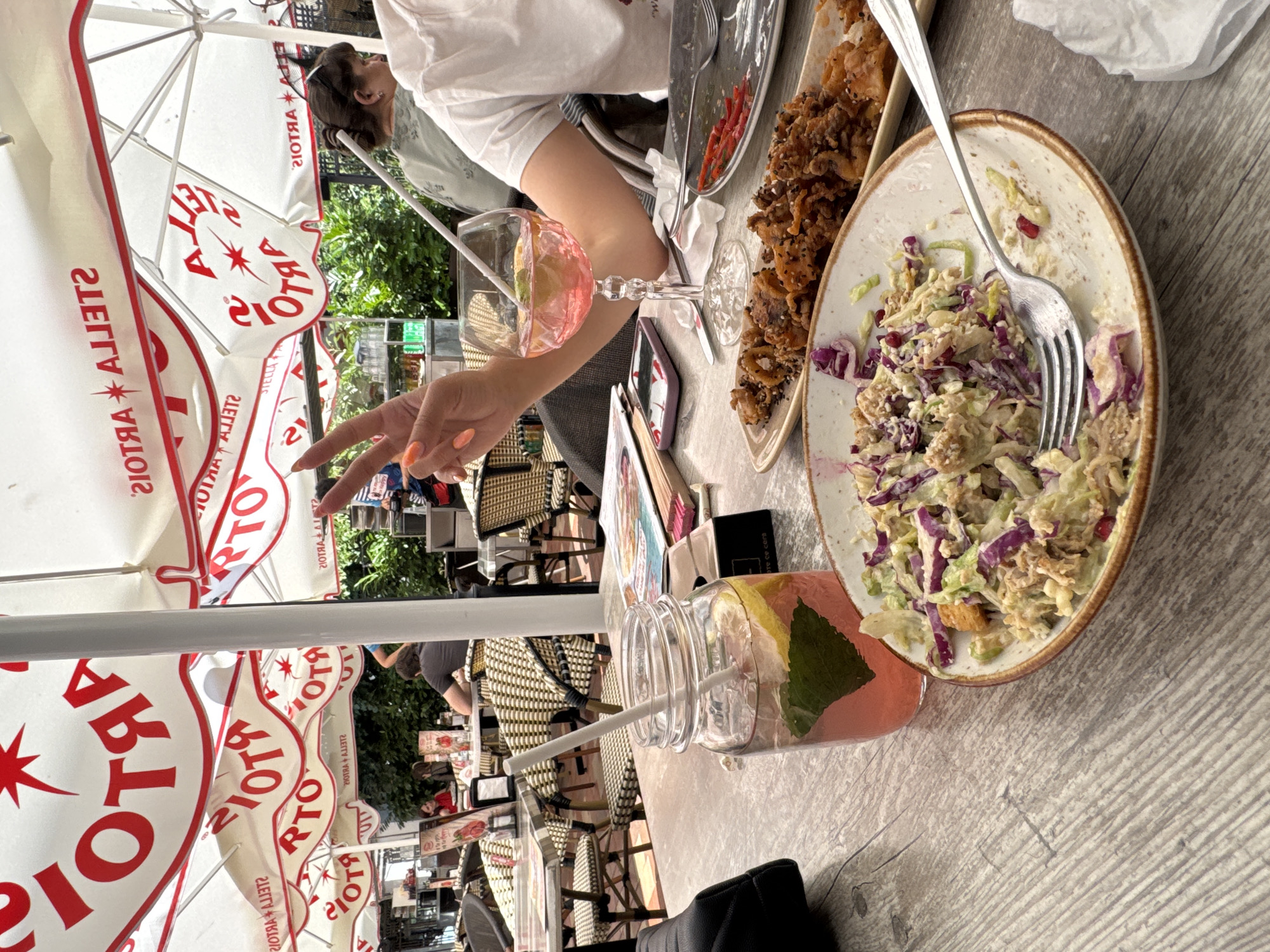 A shallow-focus outdoor shot captures a table at an outdoor restaurant, possibly in Varna, Bulgaria, under Stella Artois umbrellas. In the foreground, a partially eaten salad with red cabbage and a fork rests on a rustic wooden table. To the left, a mason jar with a pink drink, lemon slices, and mint leaves sits next to a black bag. A hand with orange-painted nails makes a peace sign over a pink cocktail in a stemmed glass. In the background, diners sit at other tables under the umbrellas, with lush greenery visible beyond.