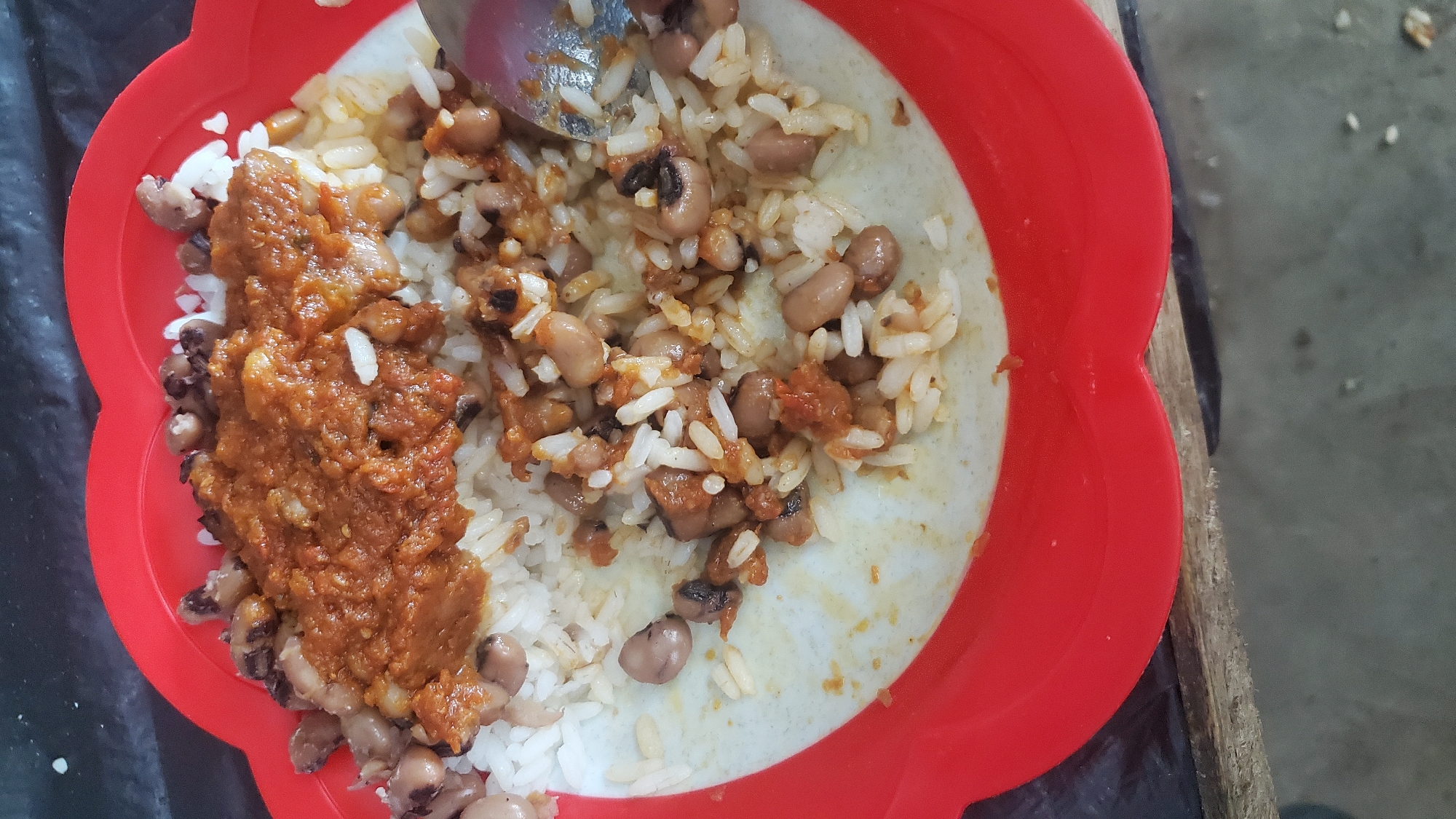 A close-up, overhead shot shows a red, scalloped plastic plate filled with white rice, black-eyed peas, and a reddish-brown sauce. The food is arranged in a mound in the center of the plate, with a portion of the rice and peas spilling over the edge onto the lighter inner rim of the plate. A silver spoon is visible on the right side of the plate, partially submerged in the food. The plate rests on a dark, textured surface, likely a table, with a sliver of a wooden surface visible at the bottom edge of the frame. The lighting is bright, casting minimal shadows. The scene appears to be an informal meal setting, possibly in a home or a casual eatery in Jalingo, Nigeria, given the typical presentation of such a dish.