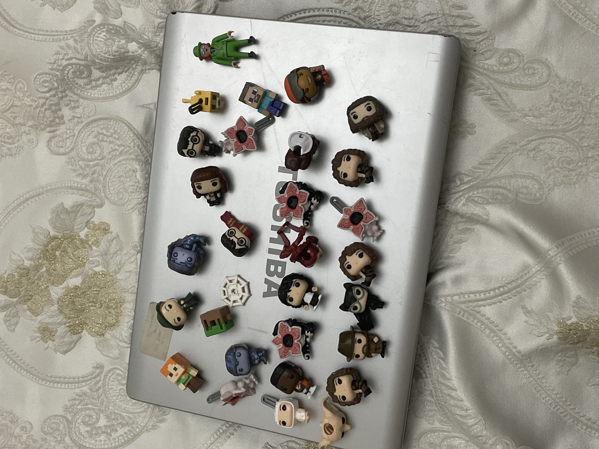 A silver Toshiba laptop lies on a light-colored, ornate fabric surface, likely a bedspread or tablecloth, which features a white patterned design with subtle gold, textured floral accents. The laptop's surface is covered with a collection of small, pop-culture-themed figures, many of which appear to be keychains or charms due to visible loops and attachments.
The figures represent various characters from different franchises. Recognizable characters include Harry Potter, Hermione Granger, Hagrid (all from Harry Potter), Steve and Alex from Minecraft, and multiple Demogorgons from Stranger Things. Other figures include a green-suited character (possibly The Riddler), a yellow blocky item, a white spiderweb, a Minecraft-style dirt block, and several other distinct but less identifiable humanoid and creature figures. Many of the figures have a stylized, large-head, small-body aesthetic, typical of Funko Pop! designs.
The figures are scattered across the laptop's lid, seemingly arranged casually for display or storage. The laptop shows visible scratches on its silver surface, indicating use. The visible text on the laptop is "TOSHIBA". There is a small, illegible sticker adhered to the bottom left of the laptop. The overall setting suggests an indoor environment. No activity is taking place other than the static display of the collectible items.