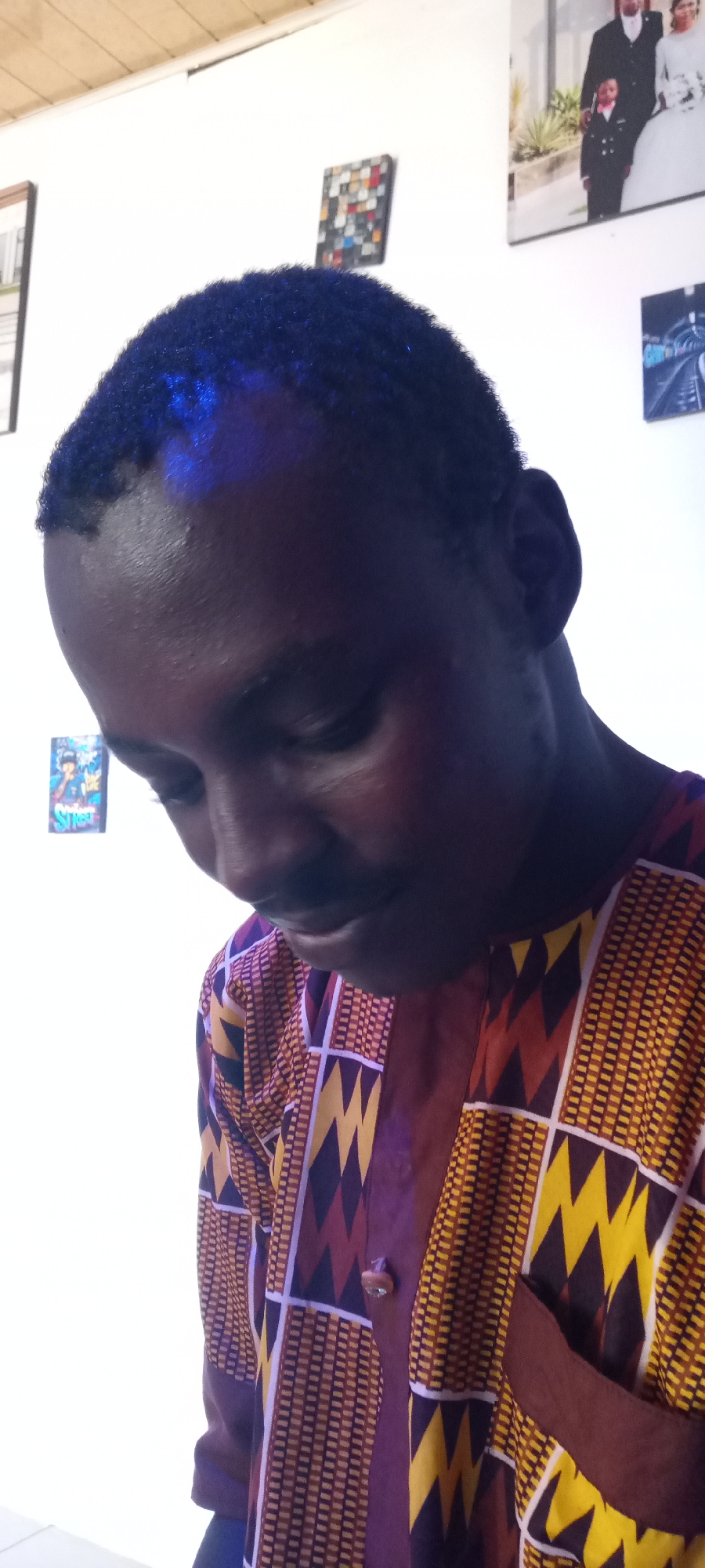 A medium shot captures a man looking downward. He is wearing a patterned shirt with yellow and brown designs and a brown pocket. The background shows a plain white wall adorned with various framed pictures. The exact location in the city of Jos, Nigeria, cannot be determined from this image.