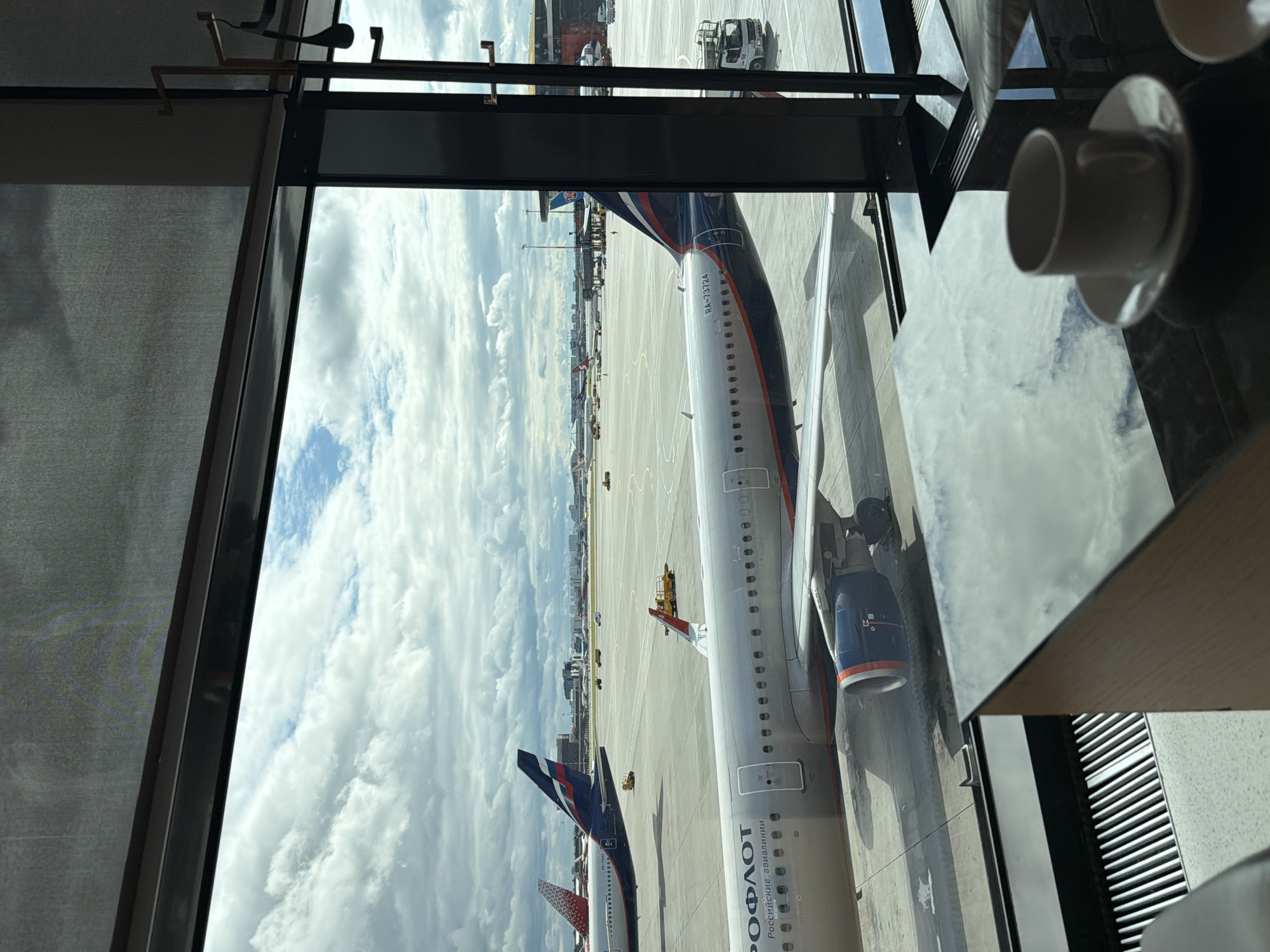 This is an eye-level shot taken from inside an airport lounge at Sheremetyevo International Airport (SVO) in Khimki, Russia, on a cloudy day. Through a large window overlooking the tarmac, several Aeroflot airplanes are visible, including one in the foreground with the registration RA-73724. The plane has its engines visible and is parked on the tarmac. In the background, other aircraft, service vehicles, and airport infrastructure are present. The interior of the lounge features a reflective black table with a white coffee cup and saucer on it, suggesting a waiting area. A heating grate is visible in the bottom left corner. The overall impression is one of transit and travel.