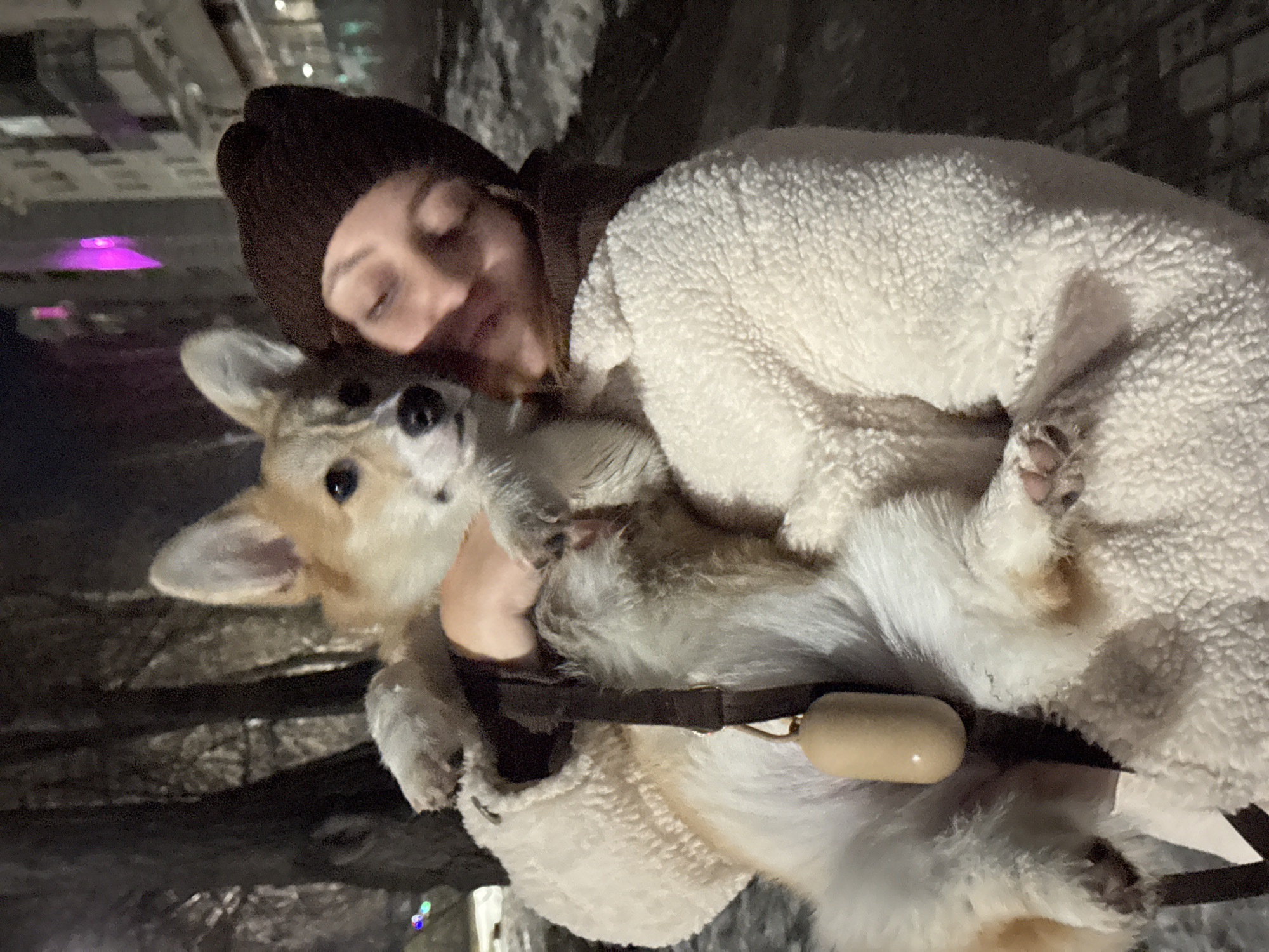 A person wearing a brown beanie and a white sherpa jacket is holding a corgi dog. The dog is wearing a brown collar with a tan-colored object attached. The location appears to be outdoors, potentially in the city of Chaikovsky, Russia, based on the prompt. The lighting suggests it may be nighttime. The person is holding the dog close, appearing to hug it. The dog is looking upward toward the camera. The environment is dark, with some ambient light sources visible.
