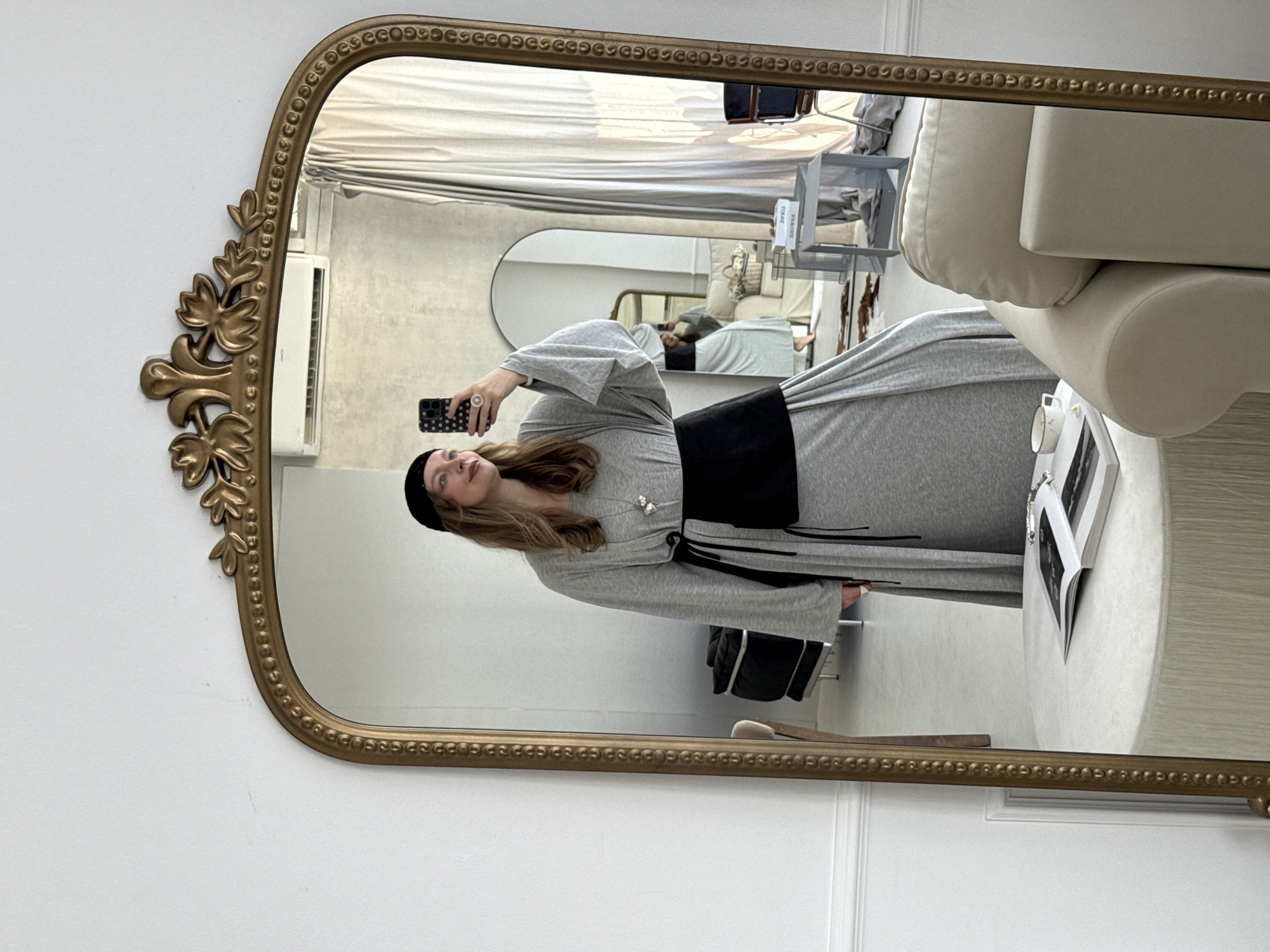 A woman is standing in front of a full-length mirror, taking a selfie. She is wearing a grey maxi dress with wide sleeves and a black sash around her waist. She also has on a black beanie and a silver pendant necklace. The mirror is ornate and gold-framed, with a decorative top. In the reflection, we can see a white wall, a light-colored couch, a round table with a book and a cup, and what appears to be a clothing rack. The overall impression is that of a well-lit indoor space, possibly a dressing room or a bedroom. The location is not specified within the image.