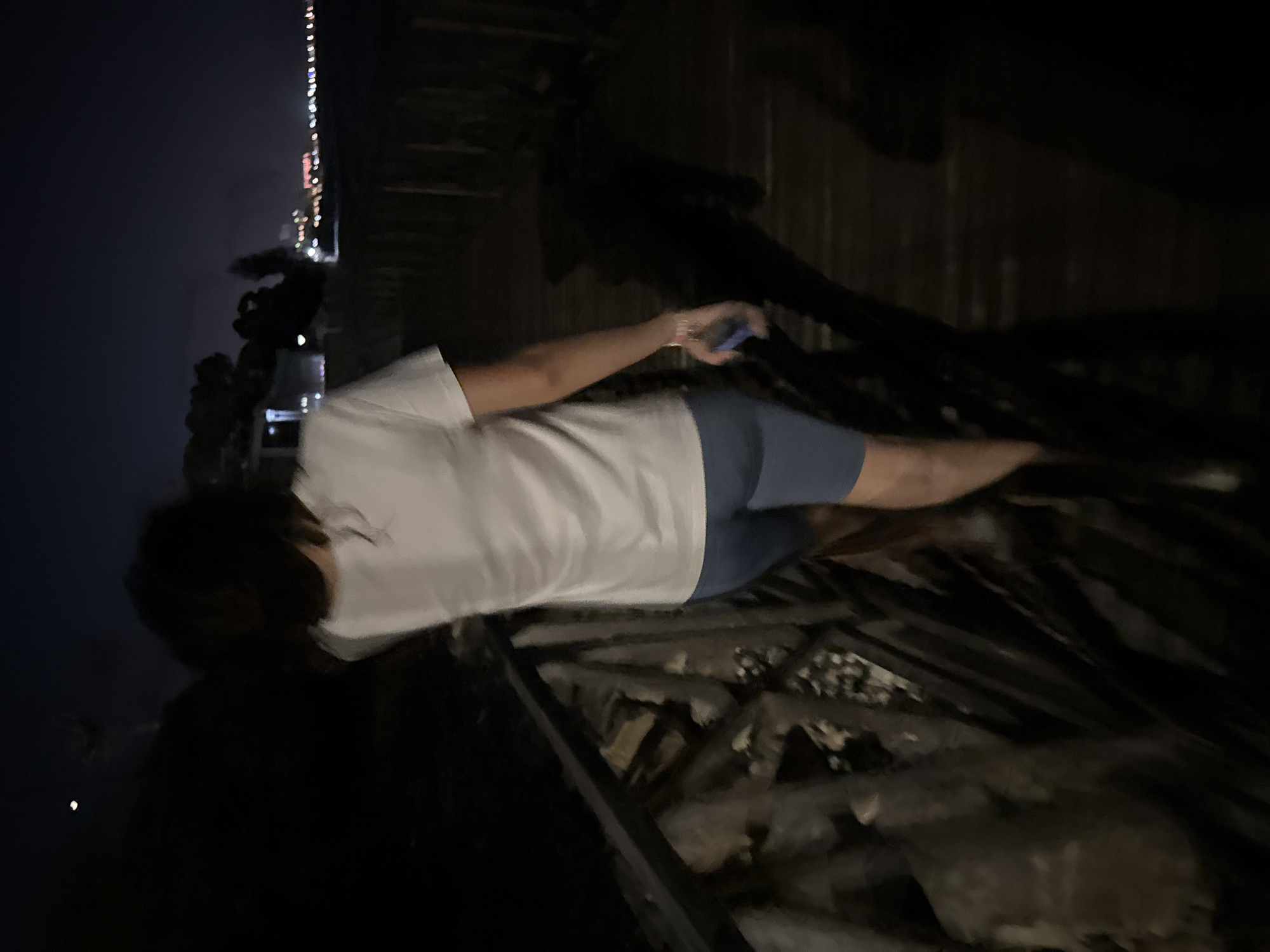 A person wearing a white t-shirt and blue shorts is walking on a wooden boardwalk at night. The boardwalk is elevated above some rocks and vegetation on the left side. In the background, there is a body of water and a city skyline with lights. The sky is dark, suggesting it is late evening or night. The overall scene appears to be in an outdoor recreational area, possibly a coastal promenade, in the Agios Tychona Community, Cyprus.
