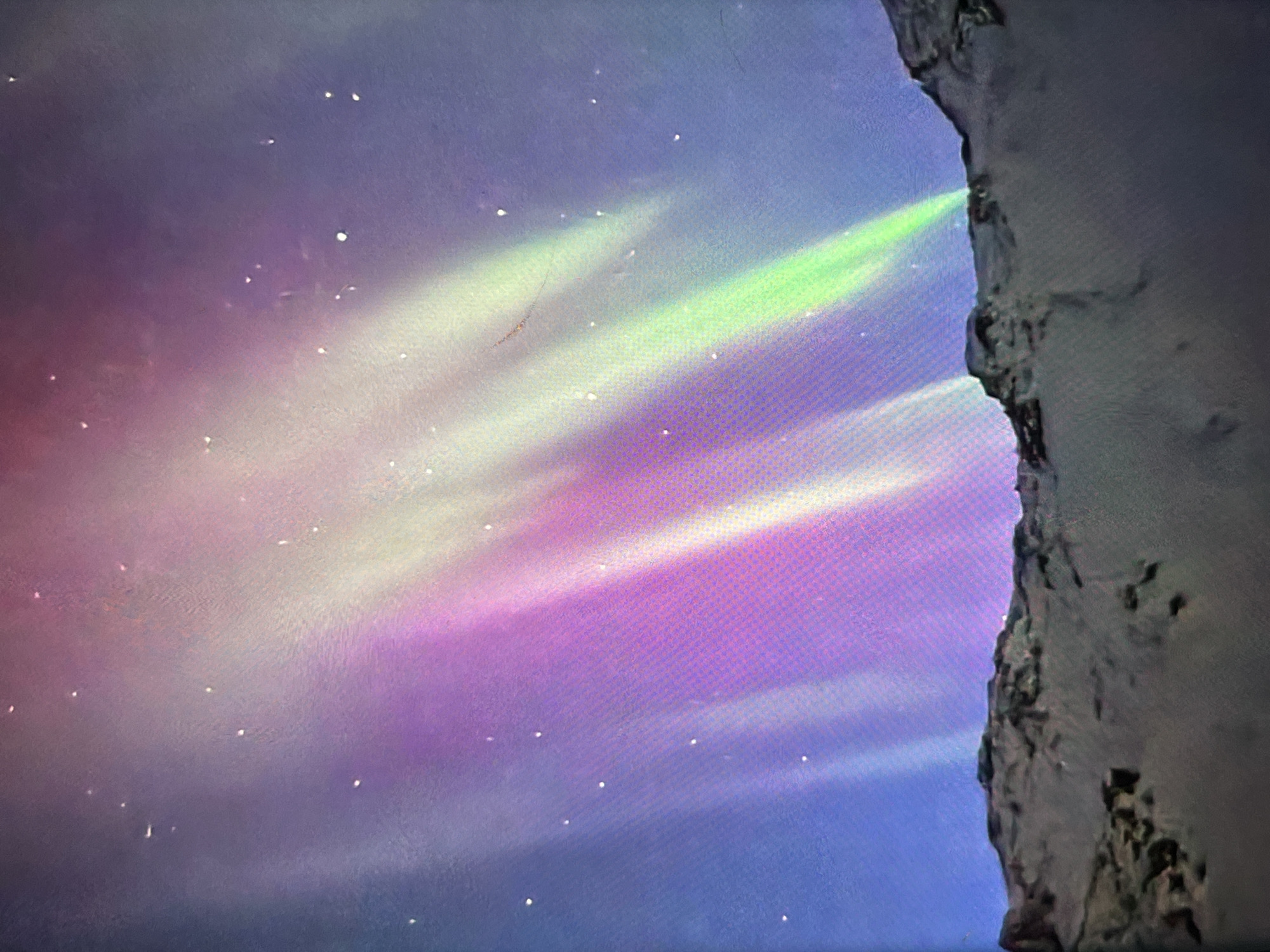 A nighttime photograph captures the aurora borealis illuminating the sky above a rugged, snow-dusted mountain range. The aurora displays vibrant streaks of purple, pink, and green light that stretch across the dark sky, interspersed with faint white dots representing stars. The foreground is dominated by the silhouette of the mountainous terrain, with visible textures of snow and rock. The overall scene is one of natural beauty and celestial phenomena.