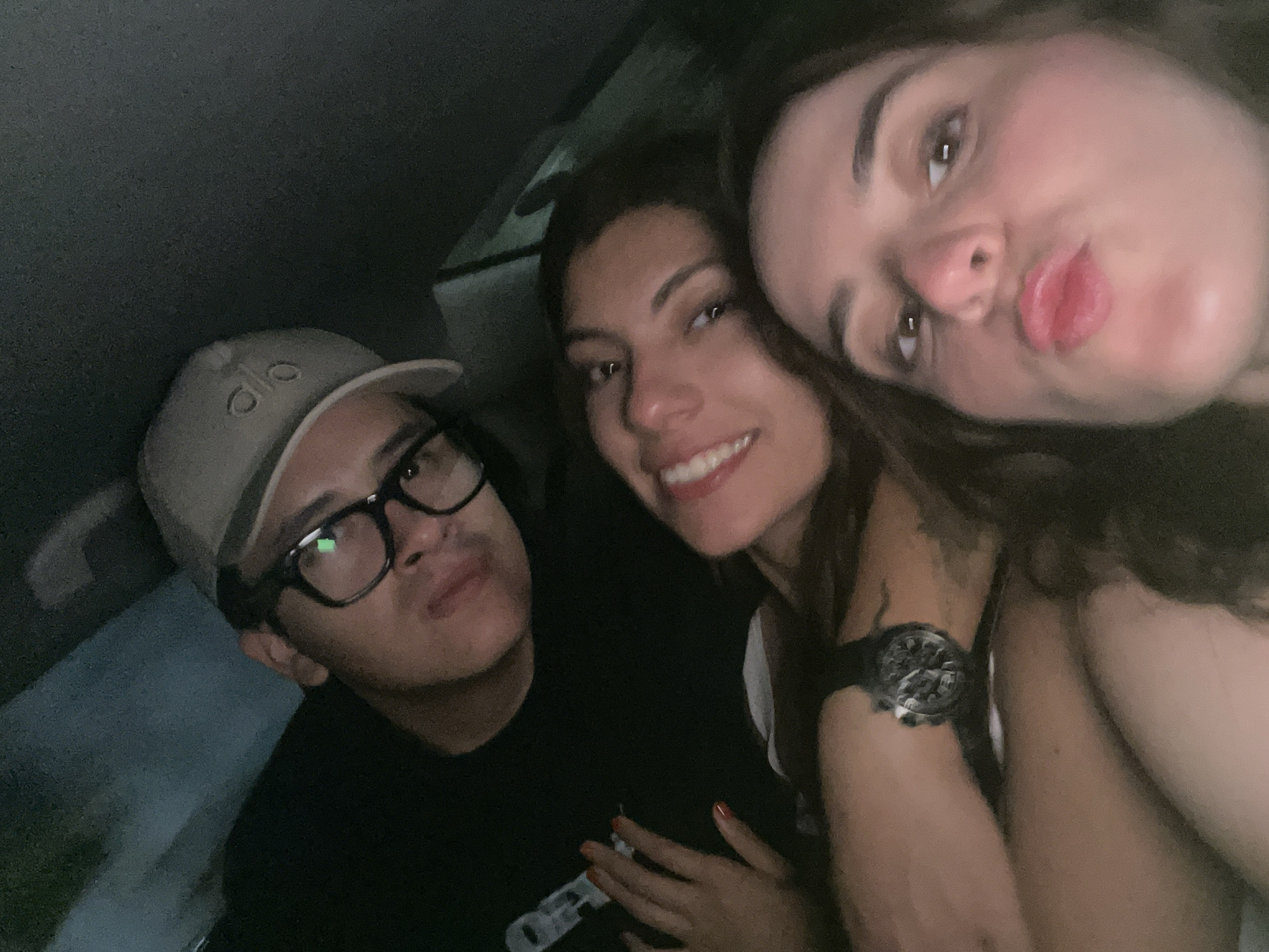 Three people are closely positioned together in what appears to be the interior of a vehicle. On the left, a person is wearing a tan-colored baseball cap with visible lettering, and thick-rimmed glasses. They are wearing a dark shirt. In the center is a person smiling with visible teeth. To the right, the third person is close to the camera, and they are pouting their lips. This person has a watch on their wrist. The scene appears dark, and the location is inferred to be within a vehicle, presumably in Brisas de Zicatela, Mexico.