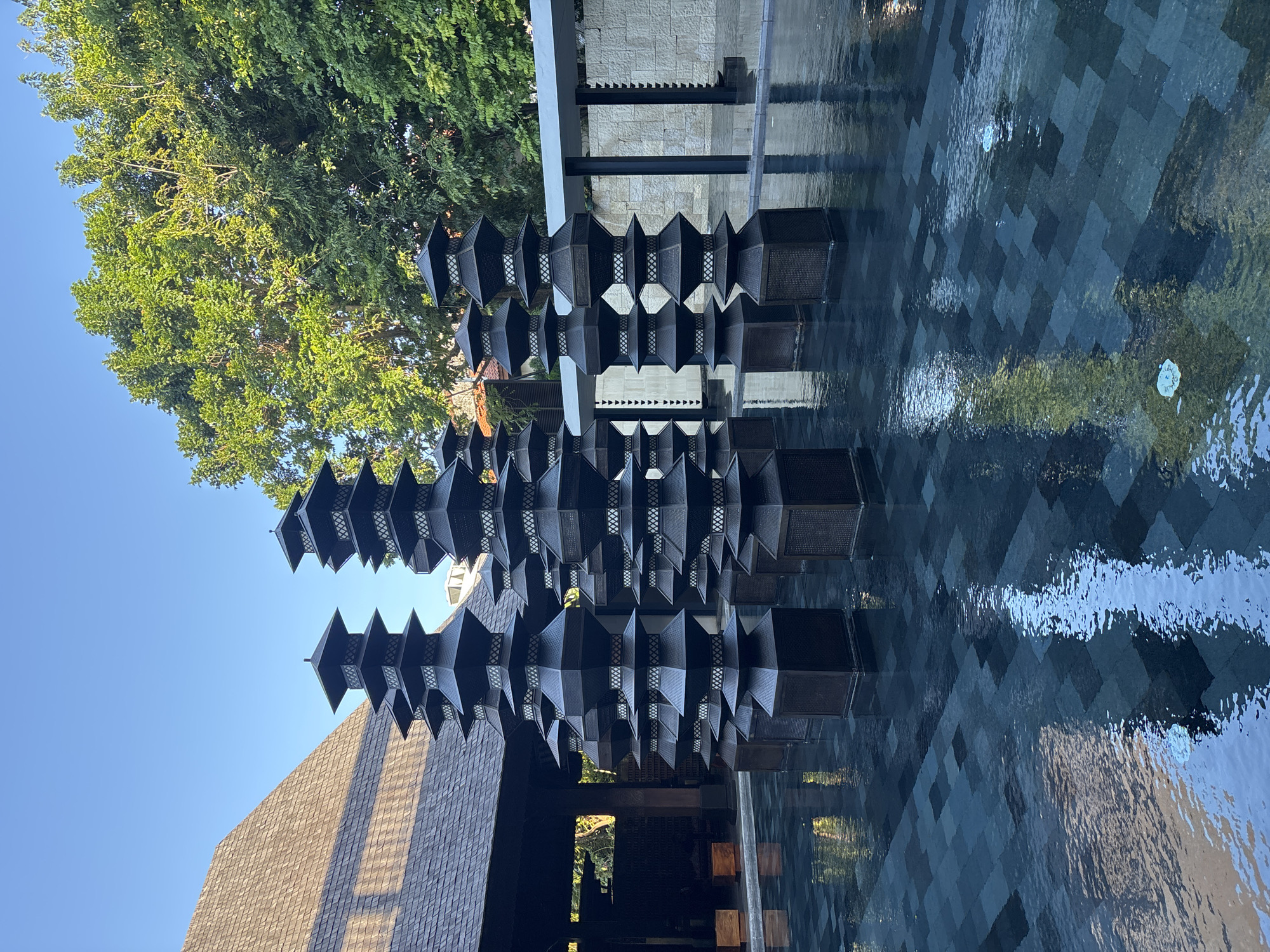 The image depicts a striking architectural scene with an emphasis on geometric forms and reflections. The primary subject is a series of tall, dark, octagonal structures that resemble tiered pagodas or lanterns, arranged in a staggered formation. These objects are made of a woven material with open latticework patterns, allowing light to pass through. They are positioned in a shallow, tiled water feature, with the dark tiles creating a grid pattern beneath the water's surface.
The water reflects the sky and the surrounding architecture, adding a dynamic and shimmering quality to the scene. To the left, a large, leafy green tree with dense foliage is visible, its branches reaching towards a clear, bright blue sky. The time of day appears to be morning or late afternoon, given the angle of the sunlight casting shadows and illuminating the scene.
Behind the decorative structures, there is a building with a stone-tiled facade and darker columns. To the far left, a portion of a building with a textured, shingled roof is visible, angled upwards towards the sky. The overall setting suggests a well-designed outdoor space, possibly a courtyard or entrance to a hotel or resort in a tropical location, given the lush greenery and bright sky. There are no people visible in the image, and the scene is serene and still, with a focus on the interplay of light, shadow, form, and reflection. The location context of Kuta, Indonesia, further supports the interpretation of a tropical, resort-like environment.