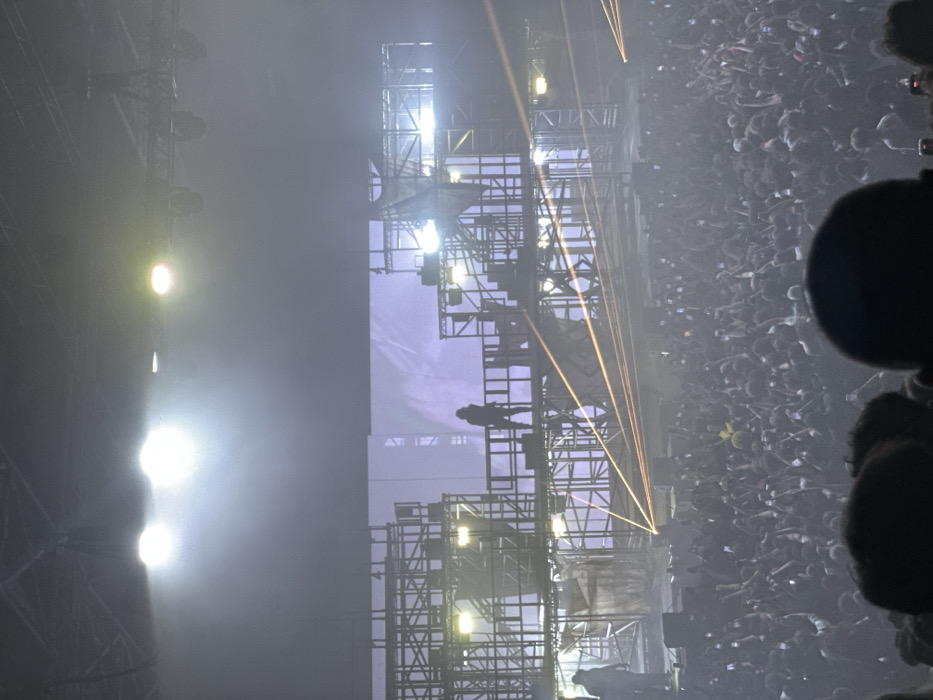 The image depicts an indoor concert scene within the Accor Arena, located at 8 Boulevard de Bercy, 75012 Paris, France. The venue is dark, with its ceiling structure and overhead rigging faintly visible.
A prominent, multi-level stage dominates the background, constructed from dark metal scaffolding, featuring platforms, stairs, and interconnected beams. Sections of the scaffolding incorporate draped or torn fabric elements, contributing to a distressed, industrial aesthetic. Behind the stage structure, a large rectangular screen displays a light purple, abstract visual.
A lone, silhouetted figure stands centrally on an elevated platform of the scaffolding, facing the audience. This figure's specific attire is obscured by backlighting, appearing as a dark form. A second, less distinct, silhouetted individual is visible on a lower platform towards the left side of the stage.
The stage is intensely illuminated by multiple bright white spotlights positioned above and behind the main performer, creating a strong backlighting effect. Numerous thin, amber-orange laser beams crisscross the stage area, cutting through a visible haze or fog that pervades the air. Additional stage lights are integrated into the scaffolding structure.
The foreground and mid-ground are filled with a dense, undifferentiated crowd of thousands of attendees, extending far back into the arena. Individual faces and features are largely indiscernible due to distance and darkness. Numerous individuals in the crowd are holding up illuminated smartphones, indicating active recording or photography of the performance. The overall scene conveys a large-scale, high-energy live entertainment event.