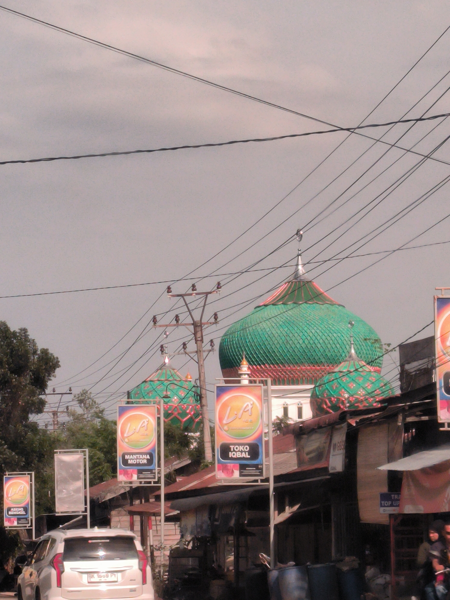 The image depicts a street scene in Aluebutuha, Indonesia, dominated by a mosque with a large green dome and smaller adjacent domes. Power lines crisscross overhead. The foreground features commercial advertisements on poles, a white SUV, and dilapidated buildings and stalls lining the street. Two individuals are visible near the stalls on the right. The scene appears to be a bustling commercial area with the mosque as a central architectural feature.