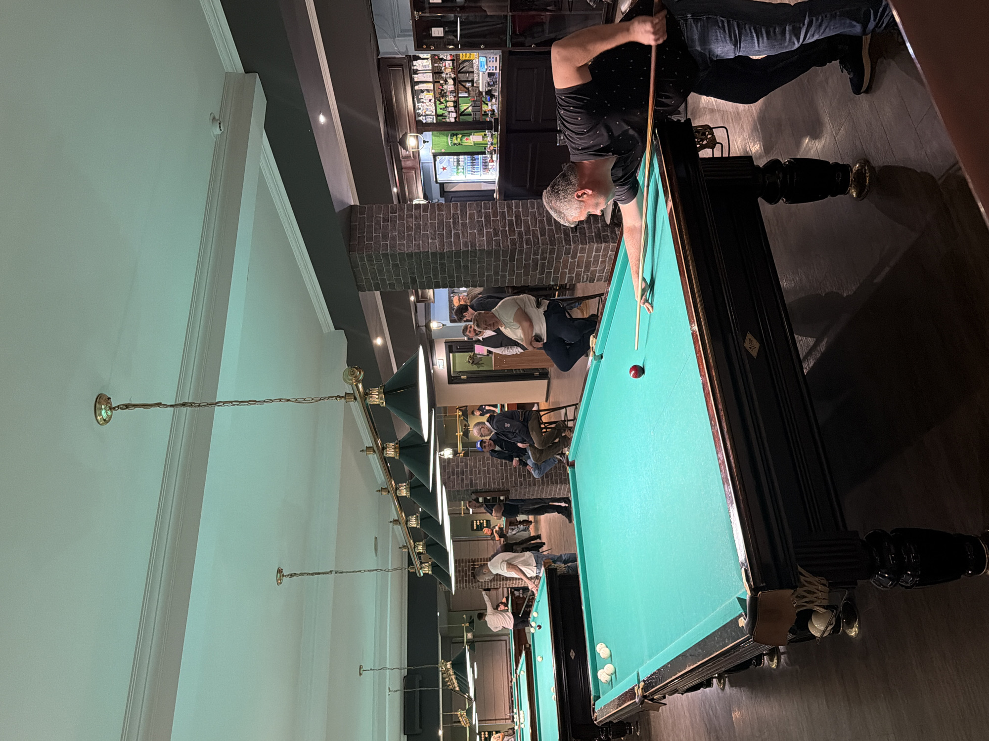 This image depicts an indoor billiard hall located in Izhevsk, Russia. In the immediate foreground on the right, a man with short gray hair, dressed in a black patterned polo shirt and blue jeans, is focused on a shot, leaning over a dark-wood billiard table with a green felt surface, holding a cue stick aimed at a single red ball. Several white billiard balls are visible clustered on the table to the left.
The establishment features multiple billiard tables, at least three of which are clearly visible, all with ornate dark wooden frames and green felt. In the mid-ground, to the left of the central player, another man in a white shirt and blue jeans stands near a billiard table with a cue stick. Further back, another man in a white shirt is leaning over a table, engaged in play. To the right, near a dark brick pillar, three men are seated on high stools, observing the activities.
The background on the right reveals a dark wooden bar counter with shelves stocked with various liquor bottles. A glass-front refrigerator, containing green-bottled beverages (with a visible Heineken star logo), stands behind the bar. Overhead, the ceiling is white with decorative molding and features multiple ornate pendant lights with dark green shades hanging above the billiard tables. A dark wooden beam with white trim runs across the ceiling, on which a smoke detector is visible. The walls are a dark gray, and the flooring is dark wood or laminate. A sign with the Cyrillic word "ВЫХОД" (EXIT) is visible above a doorway in the mid-left background.