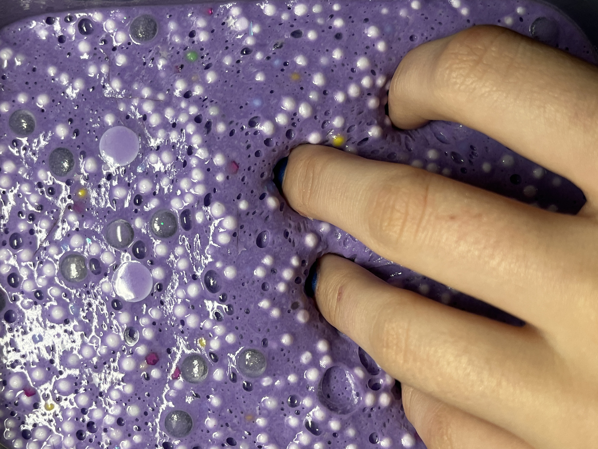 A person's hand is partially submerged in a mass of purple slime. The slime contains many small white and grey beads and some larger purple beads. The person has painted their fingernails blue. The image does not contain any information that could place it in Saint Petersburg, Russia.