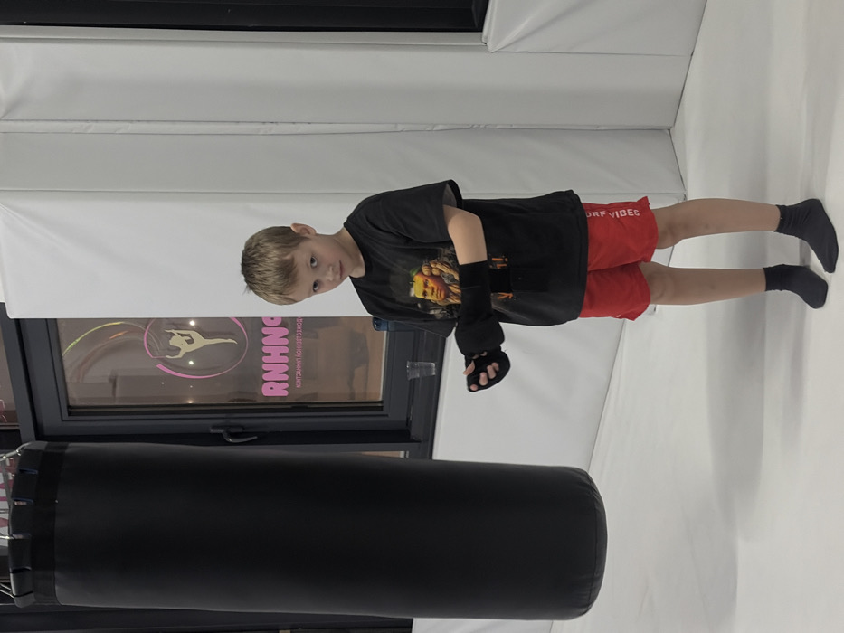 An interior image taken in Novosaratovka, Russia, depicts a young male subject standing in a combat sports or fitness facility. The subject, a boy estimated to be between five and eight years old, is positioned towards the right of the frame, looking directly at the camera with a neutral expression. He wears a black t-shirt featuring a graphic of an older male, red athletic shorts with "OFF VIBES" printed vertically on the left leg, and black socks. Black fingerless hand wraps are present on both of his hands. To the left of the subject, a large black cylindrical punching bag hangs from an unseen fixture above, partially visible. Behind the punching bag and the subject, a dark-framed window reveals a pink neon-style sign inside what appears to be an adjacent space or reflection. The sign features a stylized female figure in a dynamic pose within a circle, above the word "RNHNGA" and mirrored Russian text below, which translates to "CONTACT PHONE NUMBER." A small clear glass and a blue water bottle are visible on a ledge beneath the window. The floor is covered with a light-colored mat, and white padded walls extend along the right side and corner of the background.