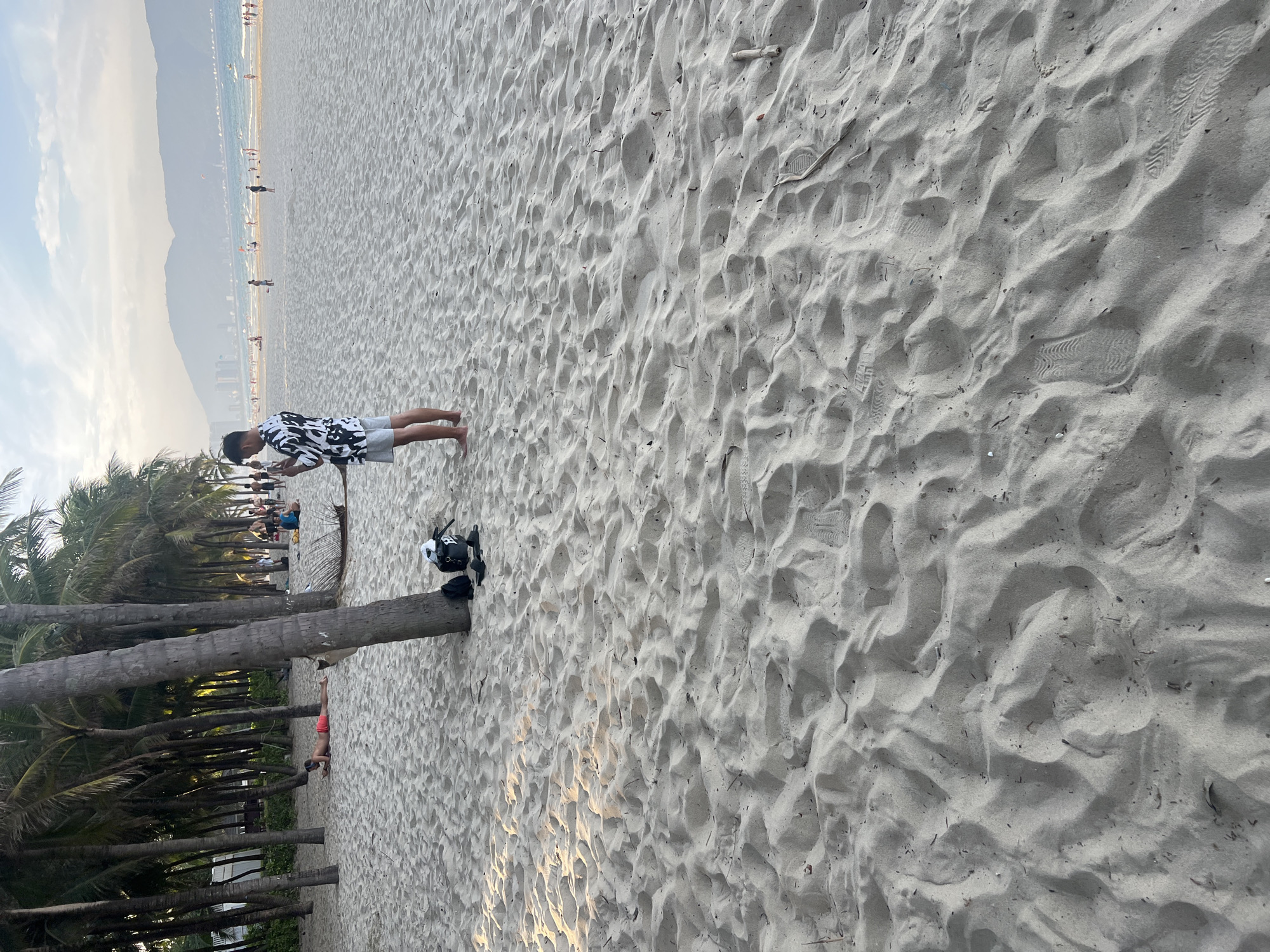 The image depicts a beach scene in Đà Nẵng, Vietnam, likely My Khe Beach. A man in a black and white patterned shirt and grey shorts stands in the sand, looking at his phone. Beside him, a black backpack and a pair of black flip-flops rest at the base of a palm tree. In the background, numerous palm trees line the beach, with other beachgoers visible in the distance. The ocean is calm, and the sky is overcast with hazy mountains on the horizon. The sand is textured with numerous footprints and wind patterns. A person in red swim trunks is lying on the sand further back, near the palm trees.