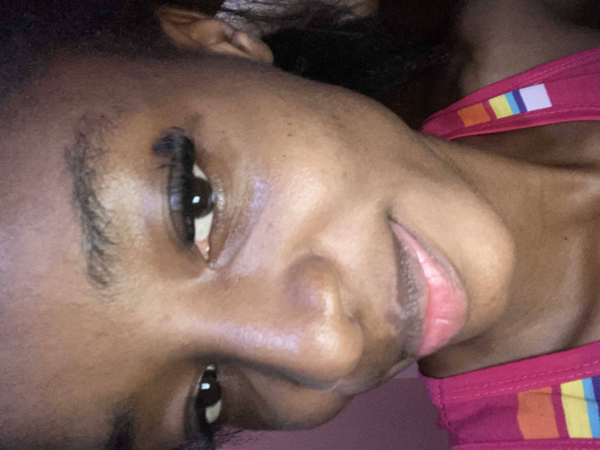 A medium close-up shot shows the face of a young Black woman who appears to be in her late teens or early twenties. She is looking slightly to her right, with a subtle smile. Her dark brown eyes have a slight glint of light reflected in them, and she is wearing dark mascara. Her eyebrows are dark and well-defined. Her nose is prominent, and her lips are full and appear to be tinted with a light pink or peachy gloss. She has a few small blemishes or moles on her face, particularly on her cheeks. Her skin tone is a rich, deep brown.
She is wearing a bright pink tank top with colorful striped detailing along the strap, featuring squares of orange, yellow, light blue, and white. The background is a solid, muted pink color, suggesting an indoor setting. The lighting is soft, with a slight glow that highlights her facial features, particularly her cheekbones and the bridge of her nose. There are no other people or objects visible in the frame besides the woman and her clothing. The image quality is somewhat grainy, which is common for photographs taken in low light conditions or with older mobile phone cameras. The overall impression is one of a casual, self-portrait style photograph.