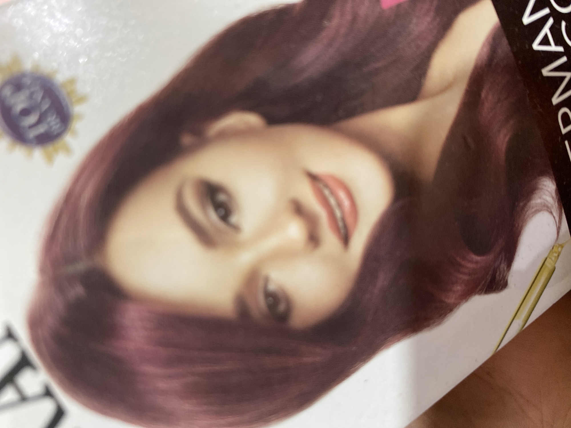 The image is a close-up shot of a woman's face and upper body, likely from a product advertisement. The woman has dark, wavy hair that appears to be dyed a deep burgundy or auburn color. She is wearing makeup, including eyeshadow and lipstick. Her expression is a slight smile. To the upper right of her head, there is a circular emblem with the word "TOP" visible. The lower portion of the image shows some text, partially obscured, that appears to say "ERMAN CO". The background is white with some subtle glitter effect. A portion of a person's hand and arm is visible on the left side of the image, and a yellow object, possibly a tube or applicator, is visible at the bottom. There is no identifiable location within the city of Ta', Indonesia in this image.