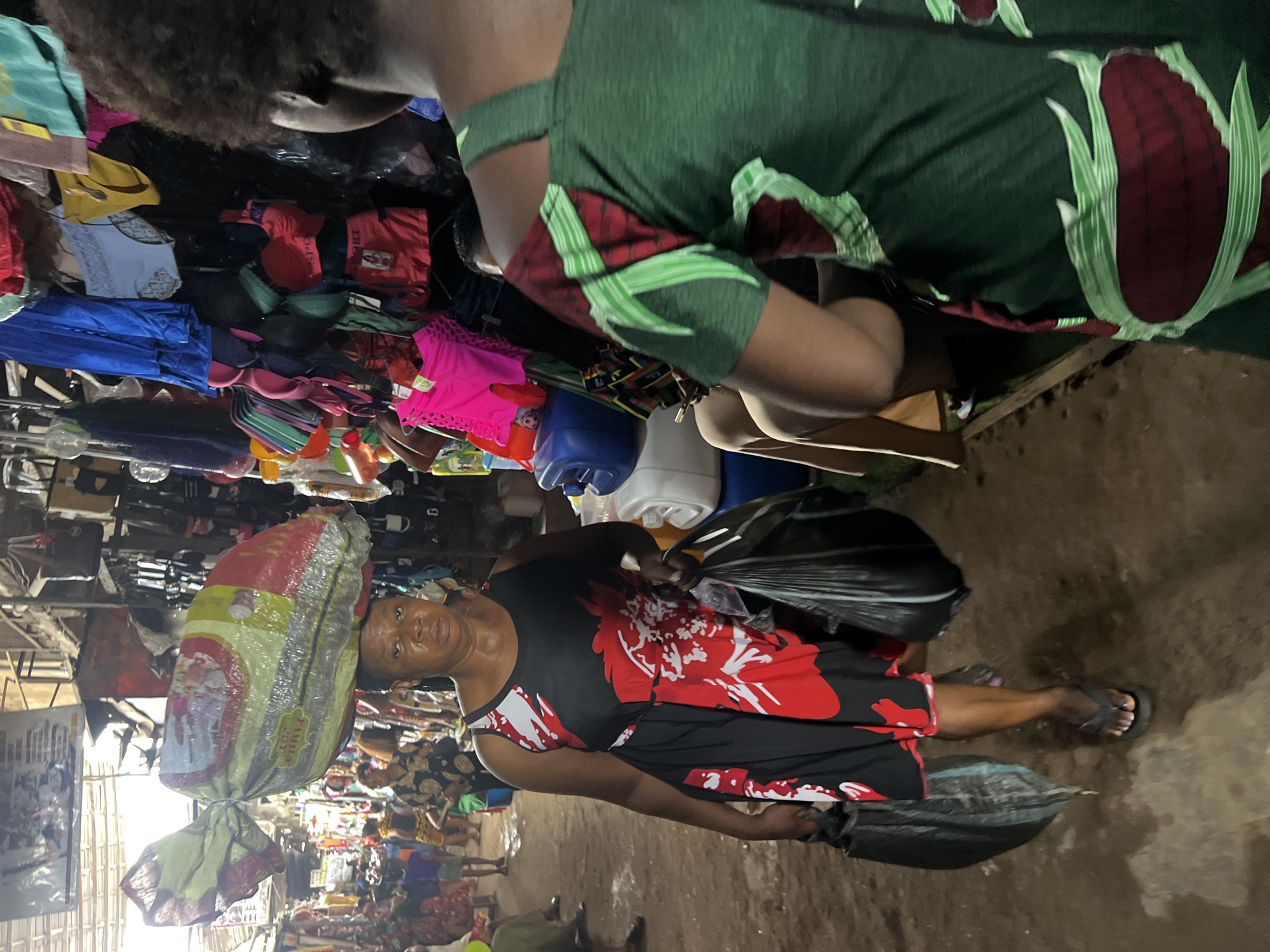 The image depicts a busy scene in Nkpor, Nigeria, likely a market. In the foreground, a woman carries a large sack on her head and holds two black plastic bags, one in each hand. She is wearing a black dress with large red floral designs and sandals. The ground is dirt and appears wet or muddy. In the background, there is a crowded area with people and various items for sale, including bras, clothing, and other goods hanging from makeshift stalls or display areas. Another person wearing green cloth with flower drawings is on top. The environment suggests a bustling marketplace with a variety of goods and people actively engaged in buying and selling.