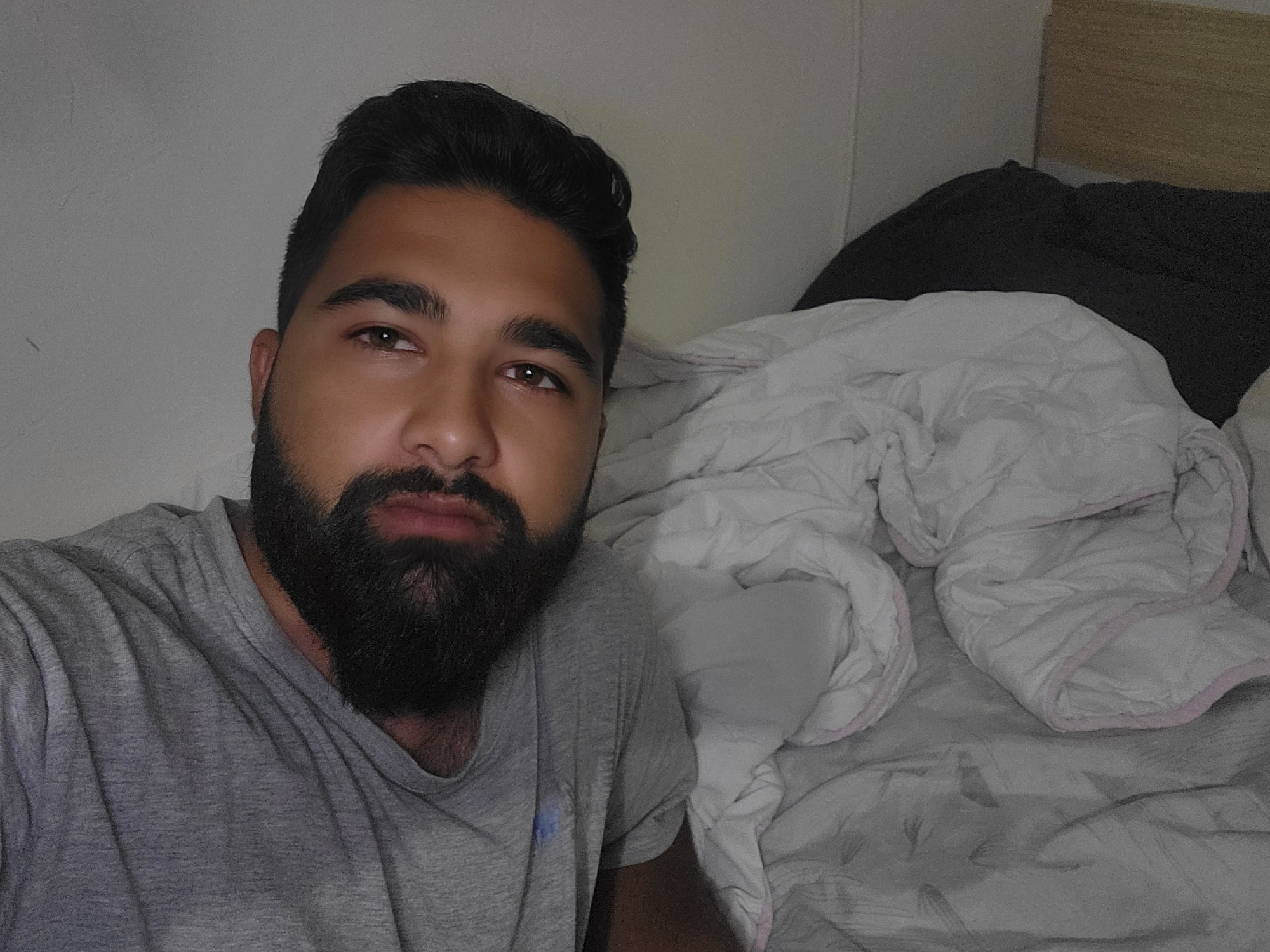 A medium, eye-level, indoor shot features a man lying in a bed. The man is in the foreground on the left side of the frame. He has dark hair, a dark beard, and is wearing a gray t-shirt. The bedsheets behind him appear white with a light pink trim. Behind the white sheets, there is a dark gray pillow and a light brown headboard. The background includes a white wall. The image does not contain sufficient information to determine if it was taken in Belovitsa, Bulgaria.