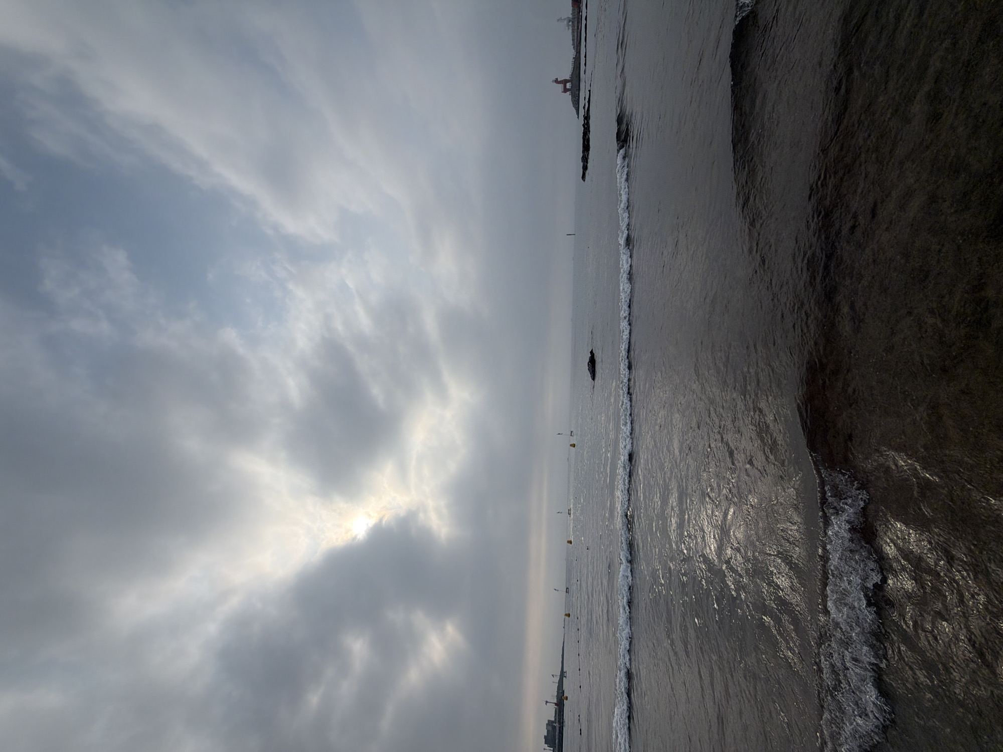 The image shows a wide shot of a serene beach scene in Jeju, South Korea, on a cloudy day. The sun is peeking through the clouds, casting a diffused light on the water. Gentle waves are lapping at the shore, and there are buoys scattered across the sea. In the distance, on the right side of the image, a distinctive red sculpture resembling a deer or horse stands on a rocky outcrop, near a lighthouse. On the left side of the image, a few buildings and cranes are visible on the coastline. The overall atmosphere is calm and tranquil, with the cloudy sky and subdued light contributing to a peaceful mood.