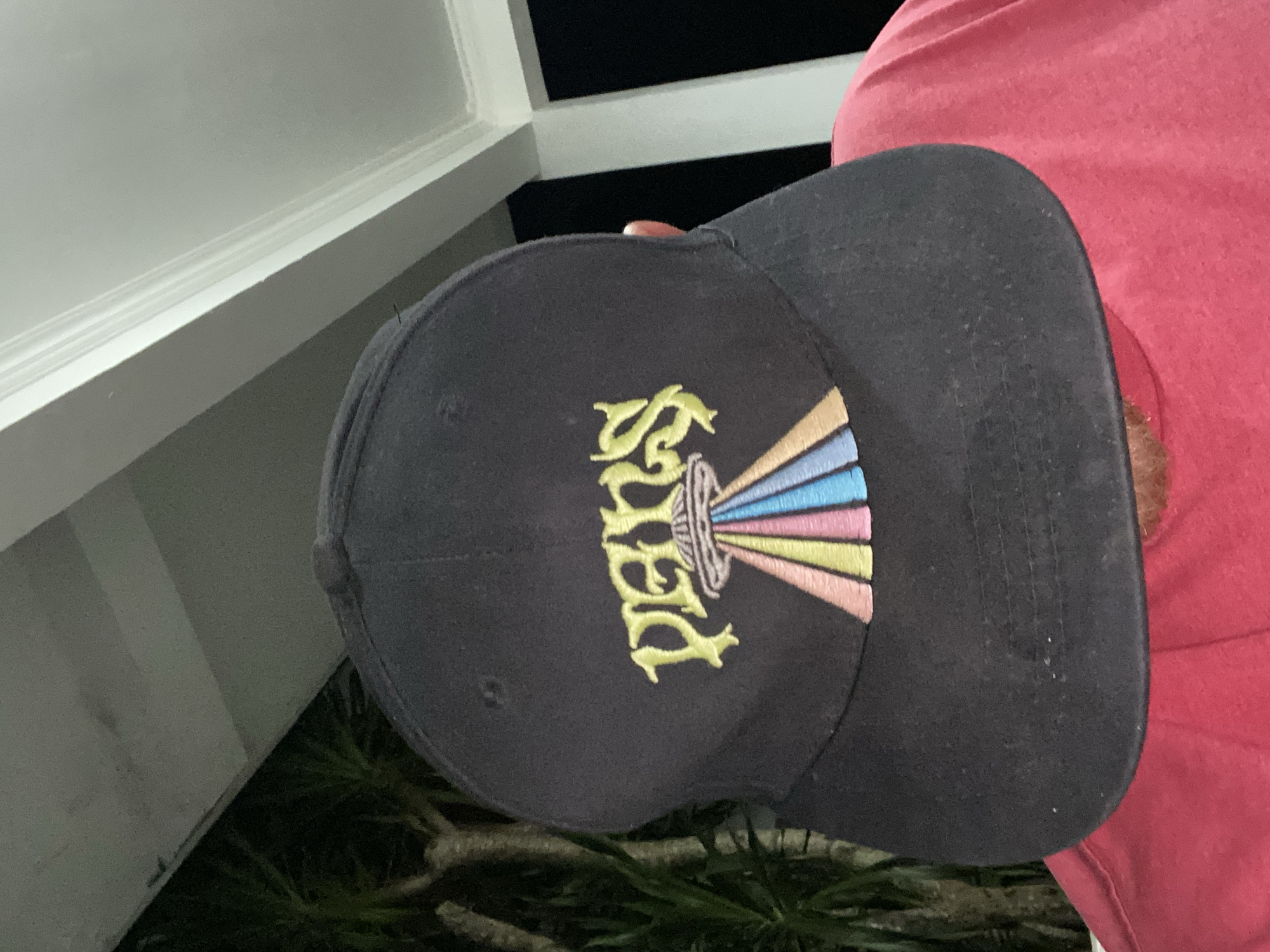 A person wearing a black baseball cap with an embroidered graphic of a UFO beaming colored light beams downward is shown from the chest up. The cap's embroidery is in a yellow, pastel color palette. The person is wearing a red t-shirt. The background shows dark foliage and white pillars, suggesting an outdoor covered area at night. The image was taken in Kuta Selatan, Indonesia.