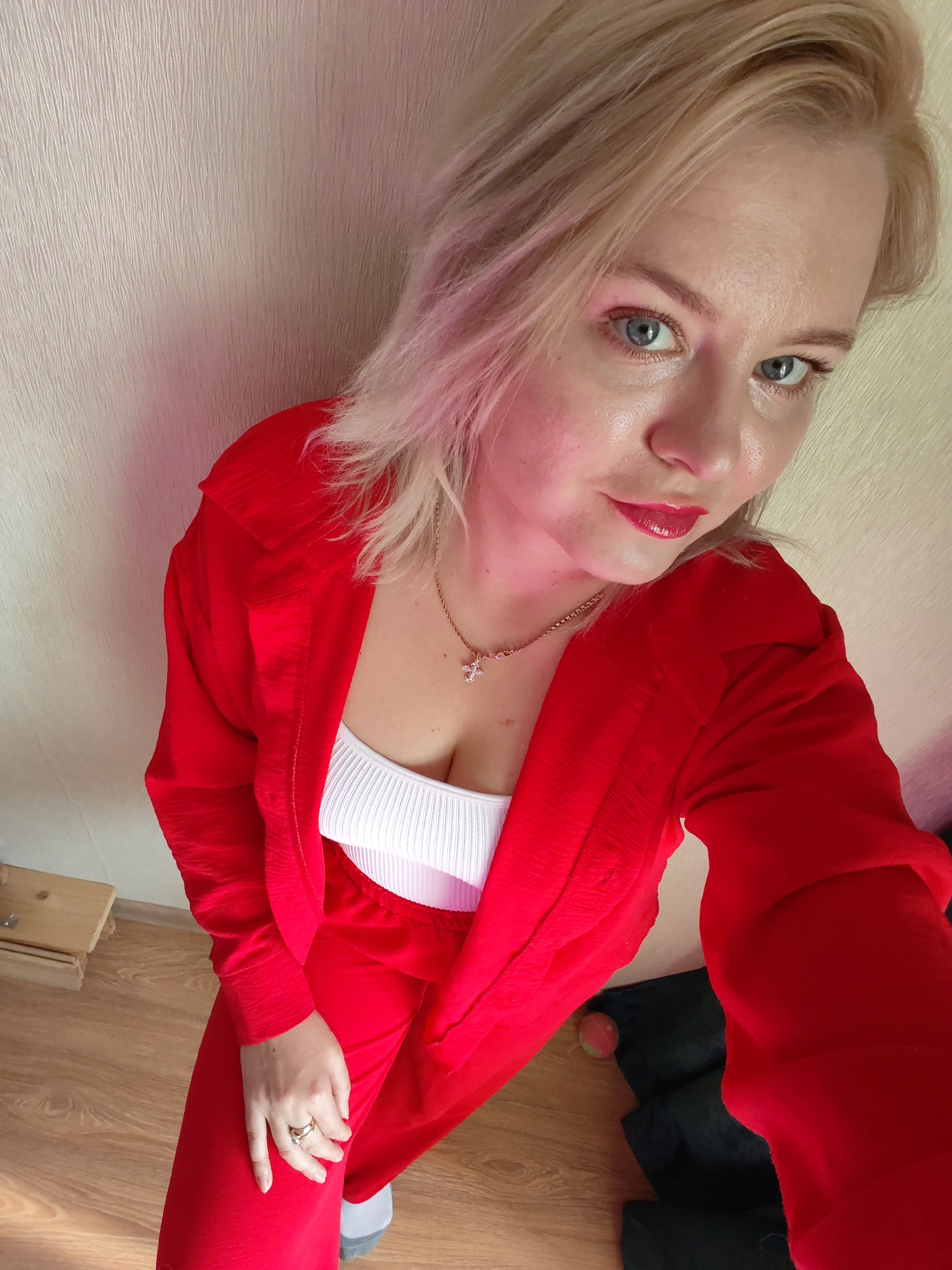 A high-angle selfie captures a blonde woman with blue eyes, wearing a red suit with a white top underneath. She is adorned with a gold necklace featuring a cross pendant and a ring on her finger. Her lips are painted with red lipstick. The background consists of a textured wall and a glimpse of the floor with some objects on it. There is no indication of the location being Izhevsk, Russia.