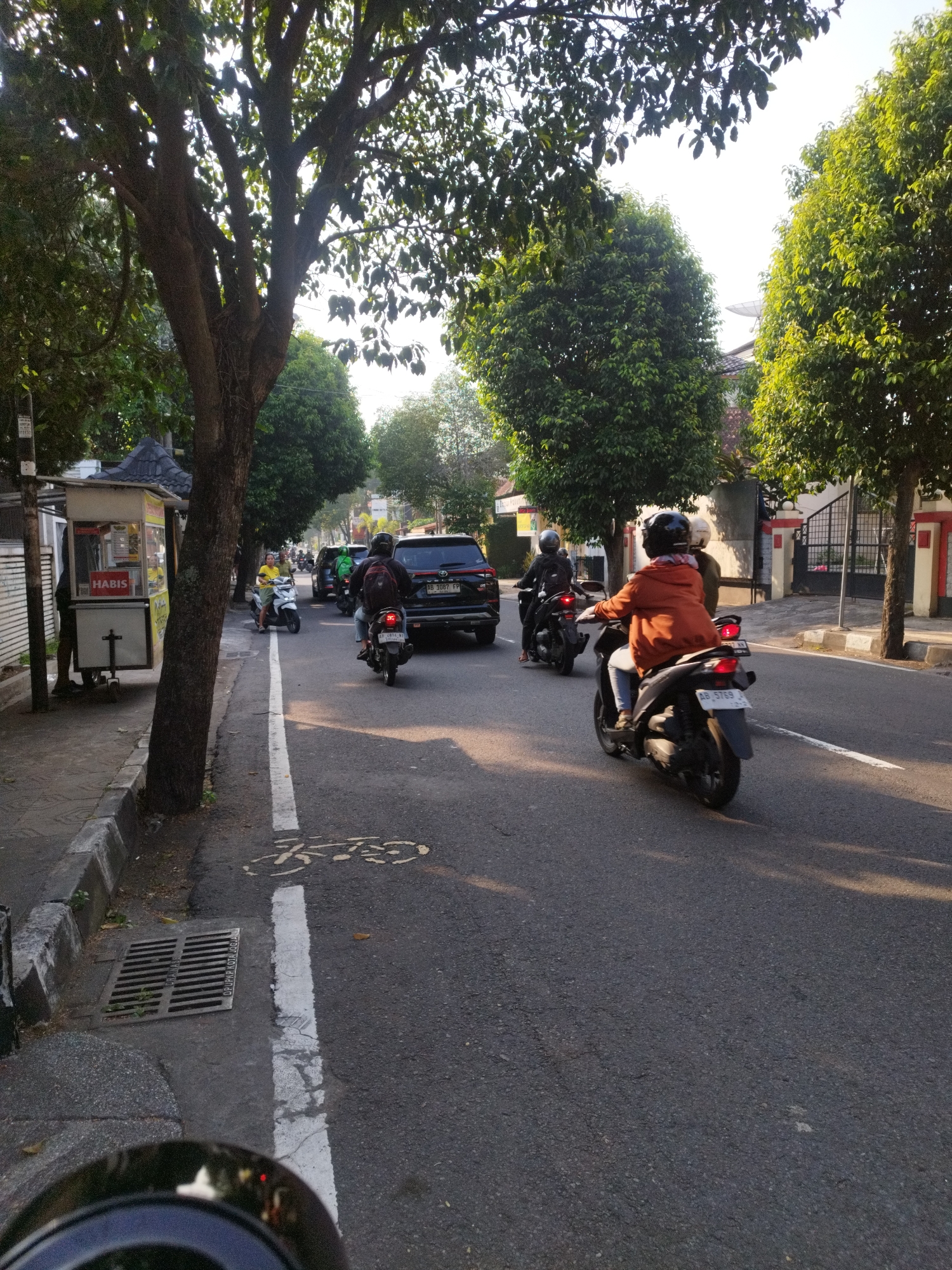 The image captures a street scene in Yogyakarta, Indonesia, likely during daytime as indicated by the lighting and shadows. The foreground is dominated by the perspective of someone riding a motorcycle, with the rear of their helmet visible at the bottom. The road surface is asphalt, marked with a white lane divider. To the left, a sidewalk is present, bordered by a curb and a drainage grate. A small food stall with a red "HABIS" sign is situated on the sidewalk, with a person standing near it.
Further down the street, traffic is moving. Several motorcycles are visible, with riders wearing helmets and jackets. One motorcycle in the mid-ground has a rider in an orange jacket. A black SUV is also in traffic. Trees line the sides of the street, providing shade and contributing to the urban environment. The background shows buildings and more trees, with a clear sky above, suggesting good weather. The overall scene depicts a typical street in a bustling Indonesian city, with a focus on vehicular traffic and everyday urban life.
