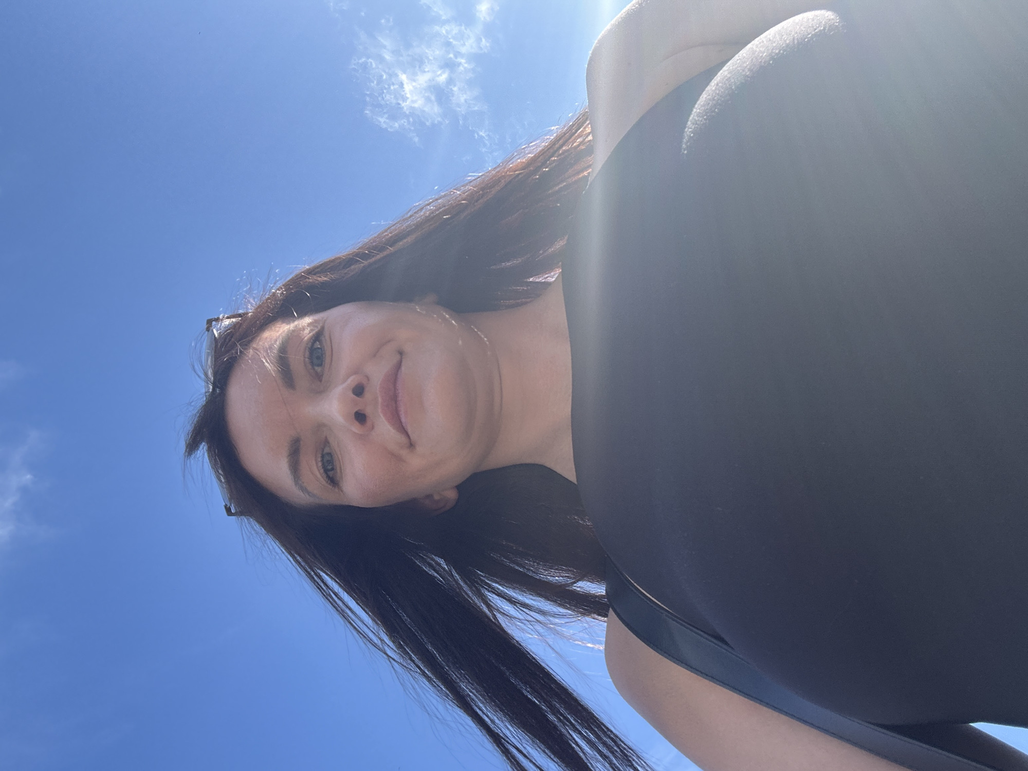 The image is a low-angle, close-up selfie of a woman with long, dark hair against a bright blue sky with scattered clouds. She is wearing a black top and sunglasses are perched on her head. The sun is casting a lens flare across the right side of the image. The woman's face is turned slightly away from the camera, and she appears to be smiling. The location is identified as Danilovka (Altai), Russia.