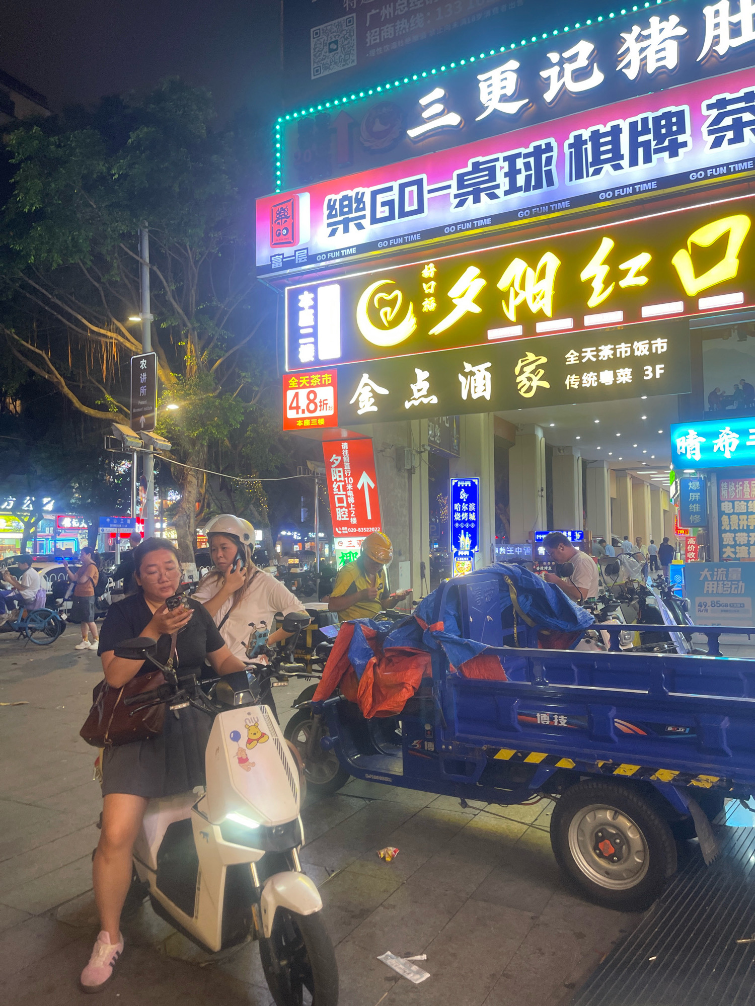 This nighttime street scene in Yuexiu District, China, captures a bustling urban environment illuminated by numerous neon signs. In the foreground, a woman wearing a black t-shirt and a grey skirt sits on a white electric scooter, looking at her phone. Next to her, another woman in a white t-shirt and helmet is talking on her phone. A blue motorized tricycle, partially covered with tarps and blankets, is parked behind them. The tricycle has yellow and black striped markings along its side and a logo with Chinese characters. In the background, several other people are visible, along with more scooters and a blue bicycle. The area is dominated by large, brightly lit signs displaying Chinese characters and advertisements, including one for "三更记猪肚鸡" (San Geng Ji Pig Stomach Chicken) and another for "乐GO-桌球棋牌" (Le GO - Billiards Chess and Cards). There are also signs for "全天茶市 饭市" (All-day Tea Market, Rice Market) and "好味道 阴红口" (Good Taste, Yin Hong Kou). A street light is visible through the branches of a large tree. The ground appears to be paved with tiles.