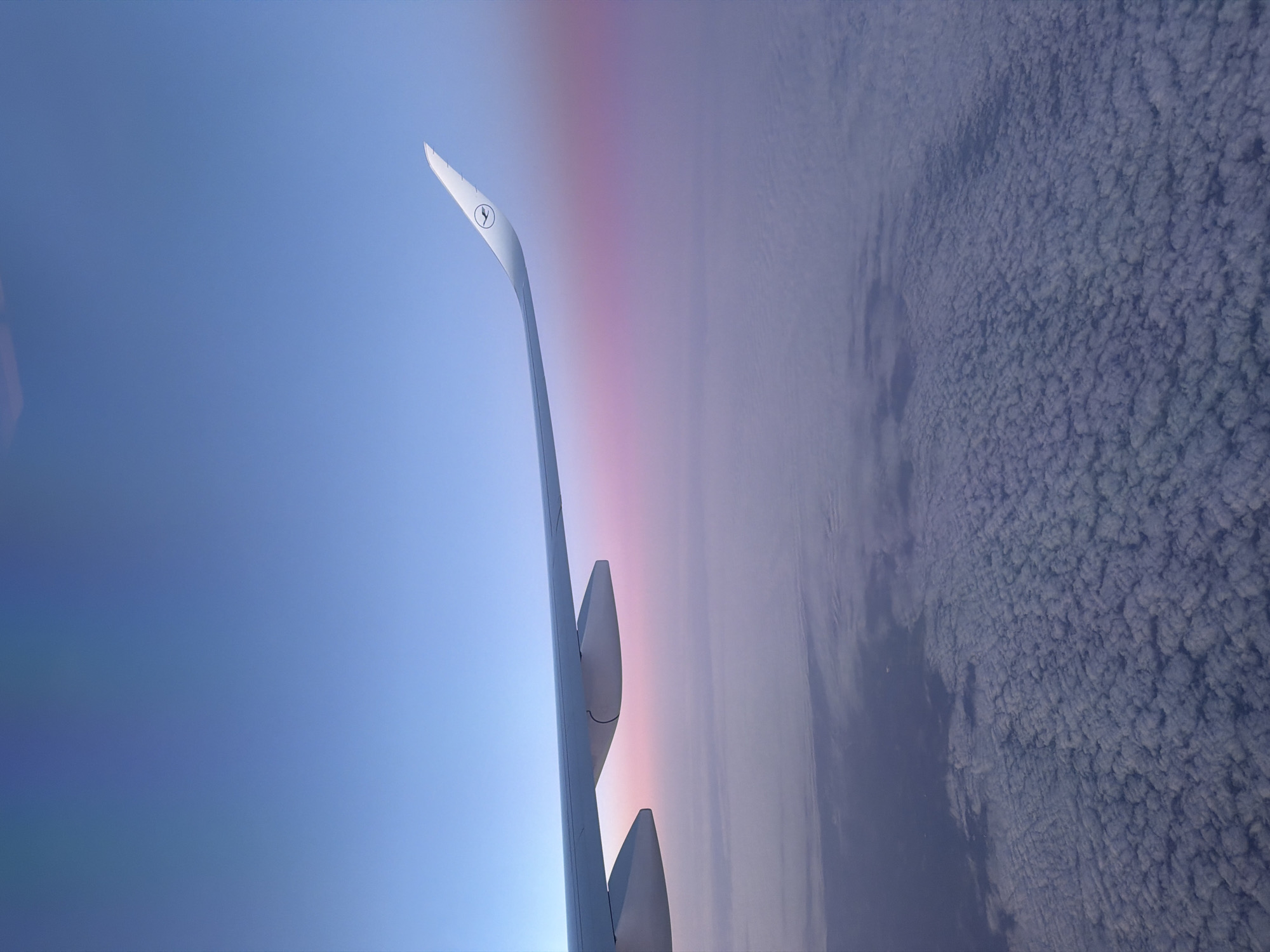 The image depicts an airplane wing, specifically the winglet with the Lufthansa logo, against a backdrop of sky and clouds. The sky transitions from a blue hue on the left to a pinkish-purple gradient near the horizon line. The clouds on the right appear as a dense, textured layer. It is not possible to determine the location in Eitting, Germany from the image.