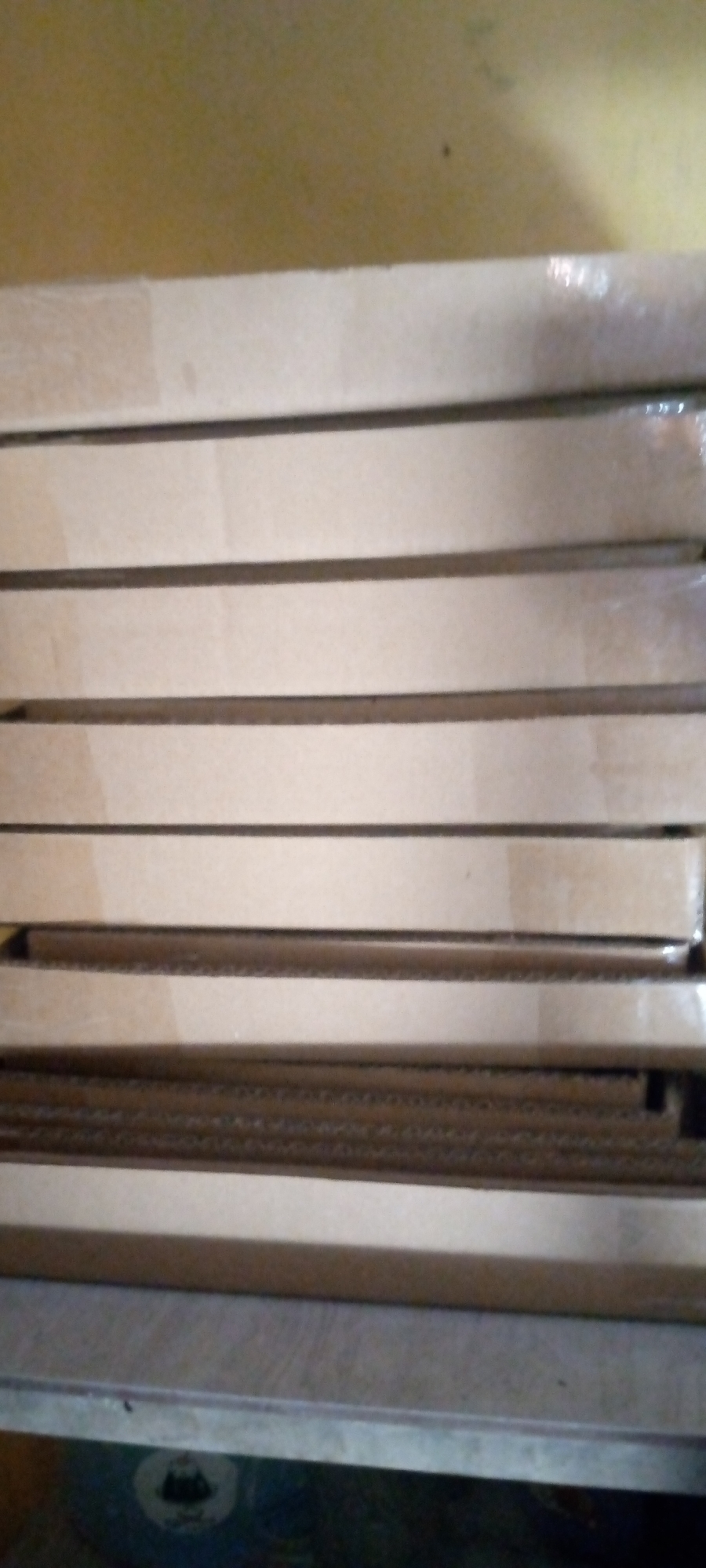 The image shows a stack of flat, rectangular objects, likely made of wood or particleboard, lying horizontally on a surface. The objects are stacked on top of each other, with a thin, dark line separating each one, suggesting they are distinct pieces. The top object is partially wrapped in clear plastic. The background is a textured, yellow-brown wall. Below the stack, a metallic or painted surface is visible, with a circular emblem featuring a black silhouette of a building or structure. The scene appears to be indoors, possibly in a storage or workshop area. The precise location in Sumberwono, Indonesia, cannot be determined from the visual information alone.
