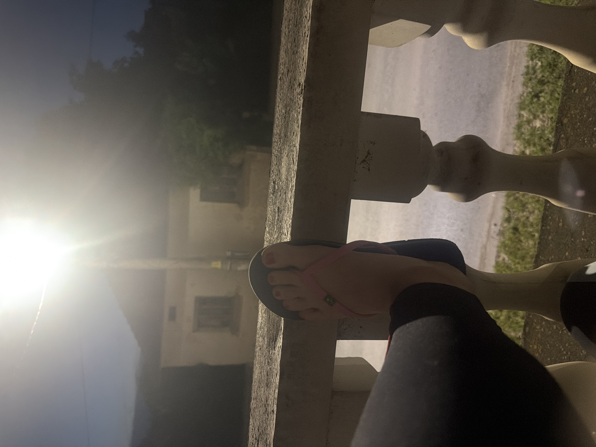 The image depicts an outdoor nighttime scene, likely captured from a balcony or porch in Iskar, Bulgaria. In the foreground, a person's bare foot, with red-painted toenails, is visible, wearing a pink-strapped flip-flop with a dark sole. The foot rests on a sturdy, light-colored railing or balustrade that shows signs of texture and wear, possibly made of concrete or painted wood. Part of the person's leg, clad in dark trousers or leggings, is also seen.
Beyond the railing, a paved path or road is discernible in the midground, with areas of green grass or weeds alongside it. Further in the background, a building with at least two rectangular windows is dimly visible. Surrounding structures and the sky are obscured by dense, dark foliage, suggesting trees or bushes.
A very bright artificial light source, likely a street lamp, emanates a strong white glow from the upper left portion of the frame, creating a significant lens flare across the image. The sky is dark, indicating it is evening or night. A small, indistinct green logo is present on the strap of the flip-flop, but its text is unreadable. The scene conveys a quiet, still moment in an outdoor setting.