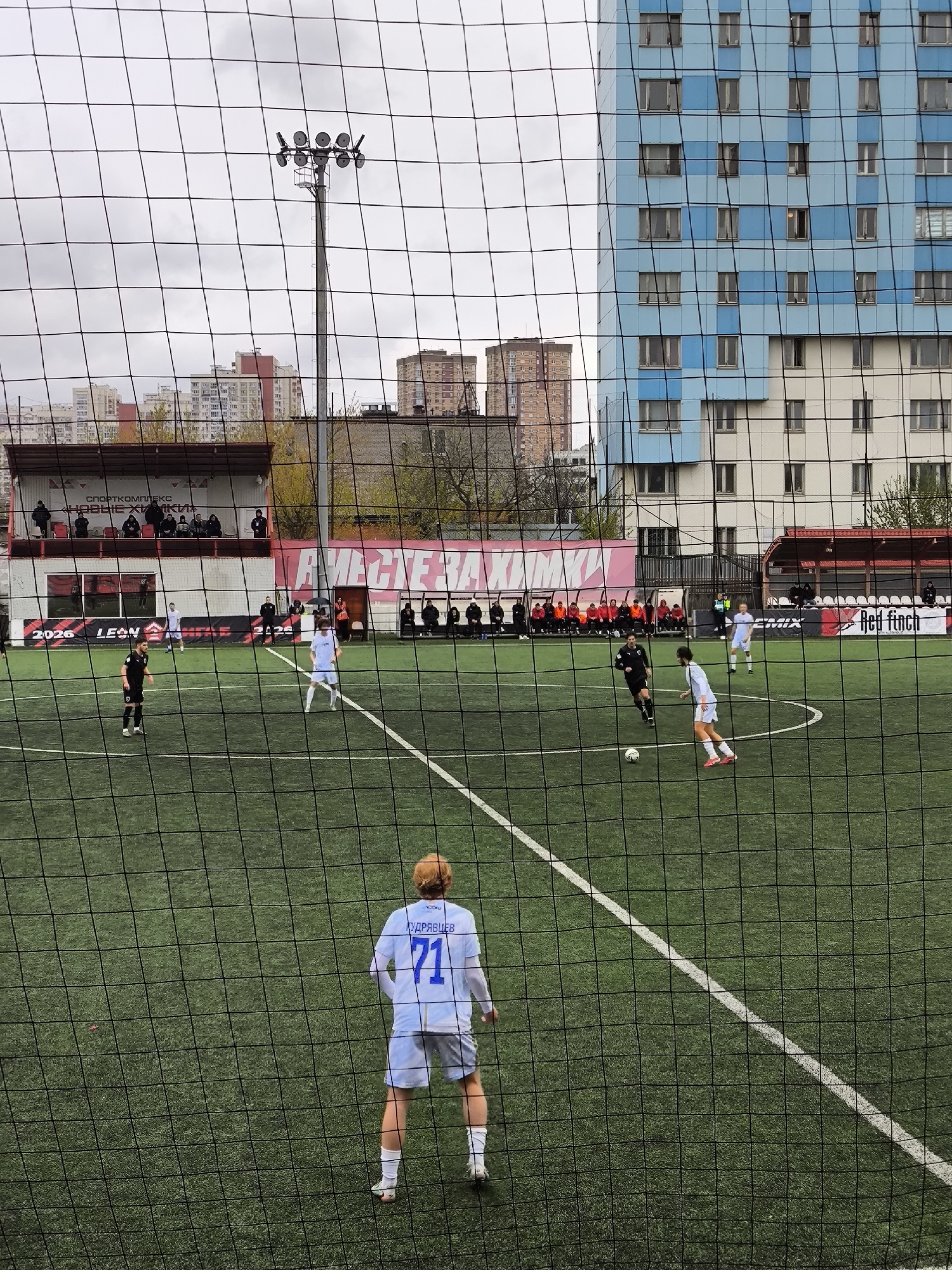 The image depicts a soccer game in progress on a green artificial turf field. The view is obstructed by a black netting. Several players wearing white and black uniforms are actively engaged in the game. Spectators are visible in the background, situated in a seating area. Banners with "ВМЕСТЕ ЗА ХИМКИ" (Together for Khimki) and sponsor logos like "Red Finch" are displayed. Buildings, including a distinct high-rise with a blue and white facade, are visible beyond the field. A light tower stands beside the field. Based on the banner, the location is Khimki, Russia.