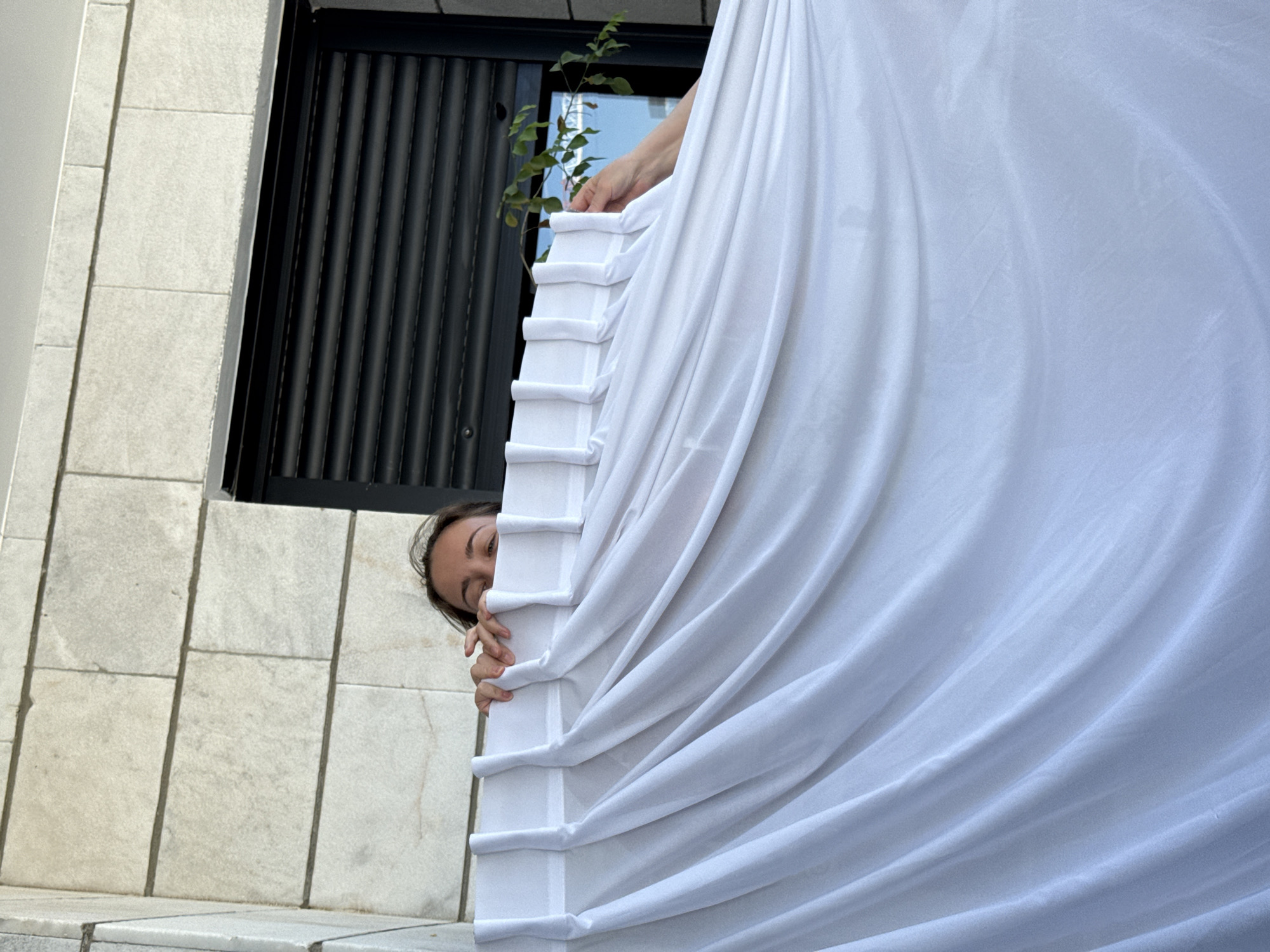 A woman peeks from behind a white curtain hanging on a balcony. She is looking towards the camera with her eyes visible above the top edge of the curtain. The curtain is draped and pleated, covering most of the frame. Behind the curtain, a building with light-colored stone tiles and a black window with rolling shutters is visible. A small green plant extends into the frame from the right. The scene appears to be outdoors, possibly on a residential balcony. This image was taken in Or Yehuda, Israel.