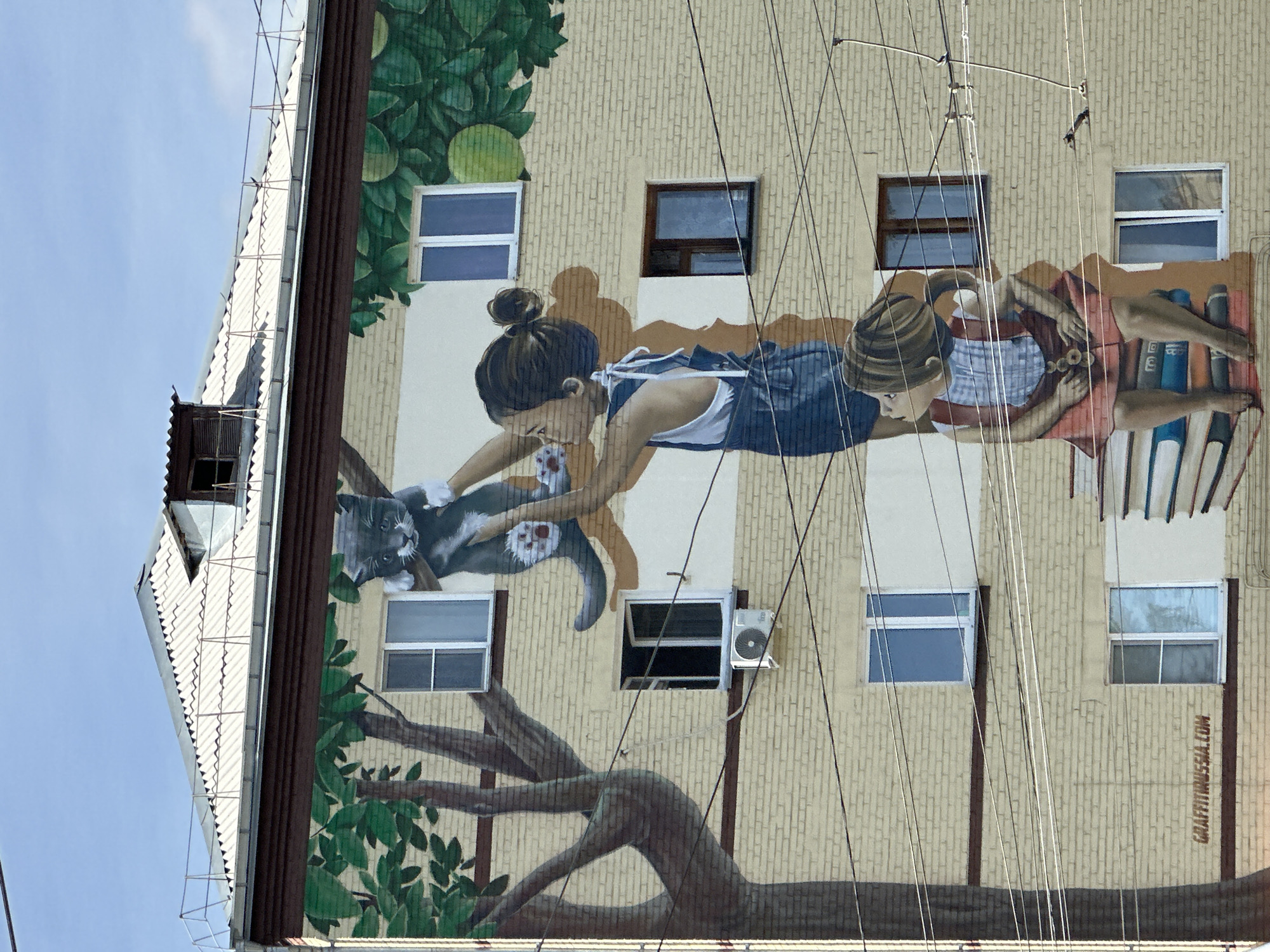 The image displays a mural painted on the side of a light yellow brick apartment building. The mural depicts two young girls and a cat. The upper girl, wearing a blue denim overall dress over a white top, is holding a grey cat up towards a tree with green leaves. The lower girl, wearing a red skirt and a blue and white checkered top, is sitting on a stack of colorful books. The scene is set against a backdrop of the building's windows, some of which are open, and an air conditioning unit is visible below one of the windows. Power lines crisscross the building. The top of the building features a sloped, corrugated metal roof with a small attic structure. A website address, "GRAFFITIRUSSIA.COM," is subtly incorporated into the mural's design near the bottom left. The sky above is a clear blue.