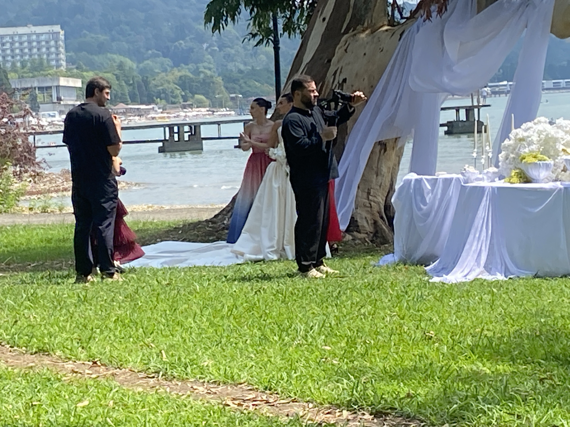 A wedding is being filmed outdoors near the sea, in Sokhumi, Abkhazia. A man in a black t-shirt and dark pants stands facing away from the camera, looking towards the sea. To his left, a woman in a long pink and blue ombre dress stands with her hands clasped. Behind her, a person in a wedding dress is partially visible. A man holding a video camera is filming the wedding party. The scene is set against a backdrop of lush green grass, a large tree draped with white fabric, and a table decorated with white flowers and candles. In the distance, there is a body of water with a pier and buildings on the shore.