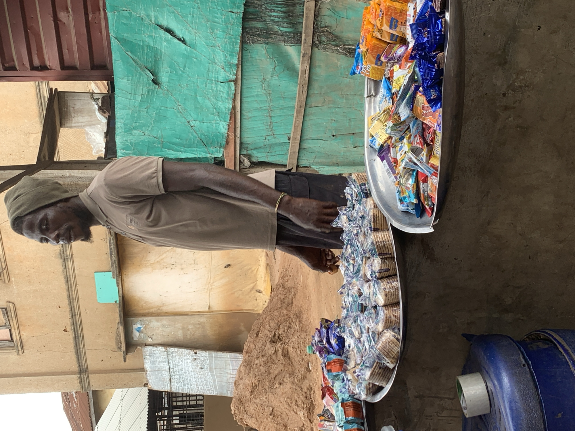 A man stands behind a stall displaying various packaged snacks and biscuits in Shagamu, Nigeria. The man wears a brown short-sleeved polo shirt and dark shorts, with a green head covering. He is positioned to the right of the frame, looking towards the left. In front of him are two large, round metal platters filled with an assortment of individually packaged snacks. One platter, closer to the viewer, contains brightly colored snack bags. The platter behind it is filled with small, stacked packages of biscuits. To the left of the platters, a blue plastic container is visible, with a roll of tape on top. The stall is set up against a building with textured walls and a corrugated metal roof. Behind the stall, a large mound of reddish-brown sand is present. The background also features green tarpaulins stretched across wooden frames. The scene appears to be an outdoor market stall.