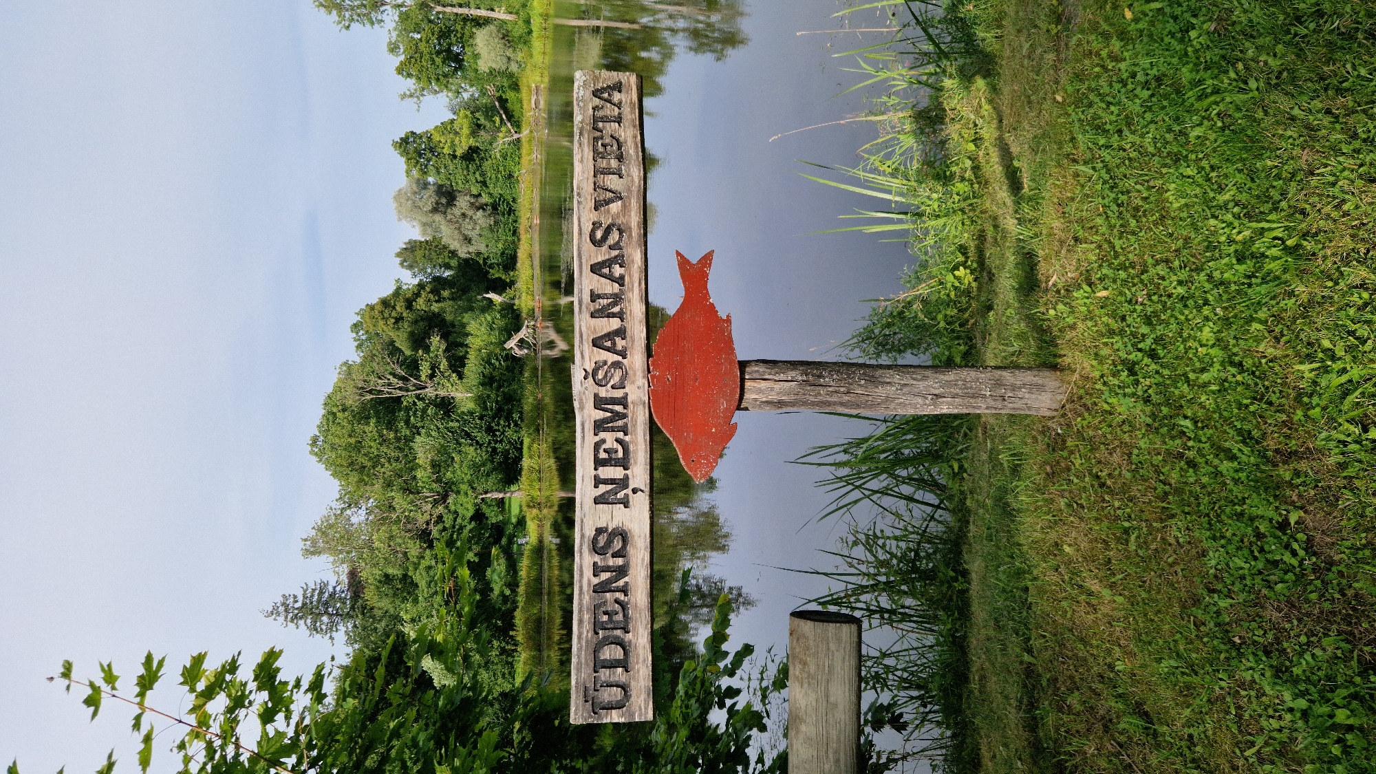 A wooden sign with the inscription "ŪDENS NEMŠANAS VIETA" (meaning "Water Intake Place") is positioned in front of a calm body of water, likely a pond or lake, surrounded by lush green trees and vegetation. The sign is mounted on a weathered wooden post, and below it, a red, silhouette-style cutout of a fish is attached to the post, indicating a fishing-related location. The environment appears serene and natural, suggesting it is located in a park or rural area. The water reflects the surrounding greenery and the sky, which is a pale blue with scattered clouds. The foreground consists of mown grass, with some taller reeds and other plants growing near the water's edge. The overall impression is of a tranquil, outdoor setting in Kazdanga, Latvia, marked for a specific activity related to the water. There are no people or animals visible in the image.