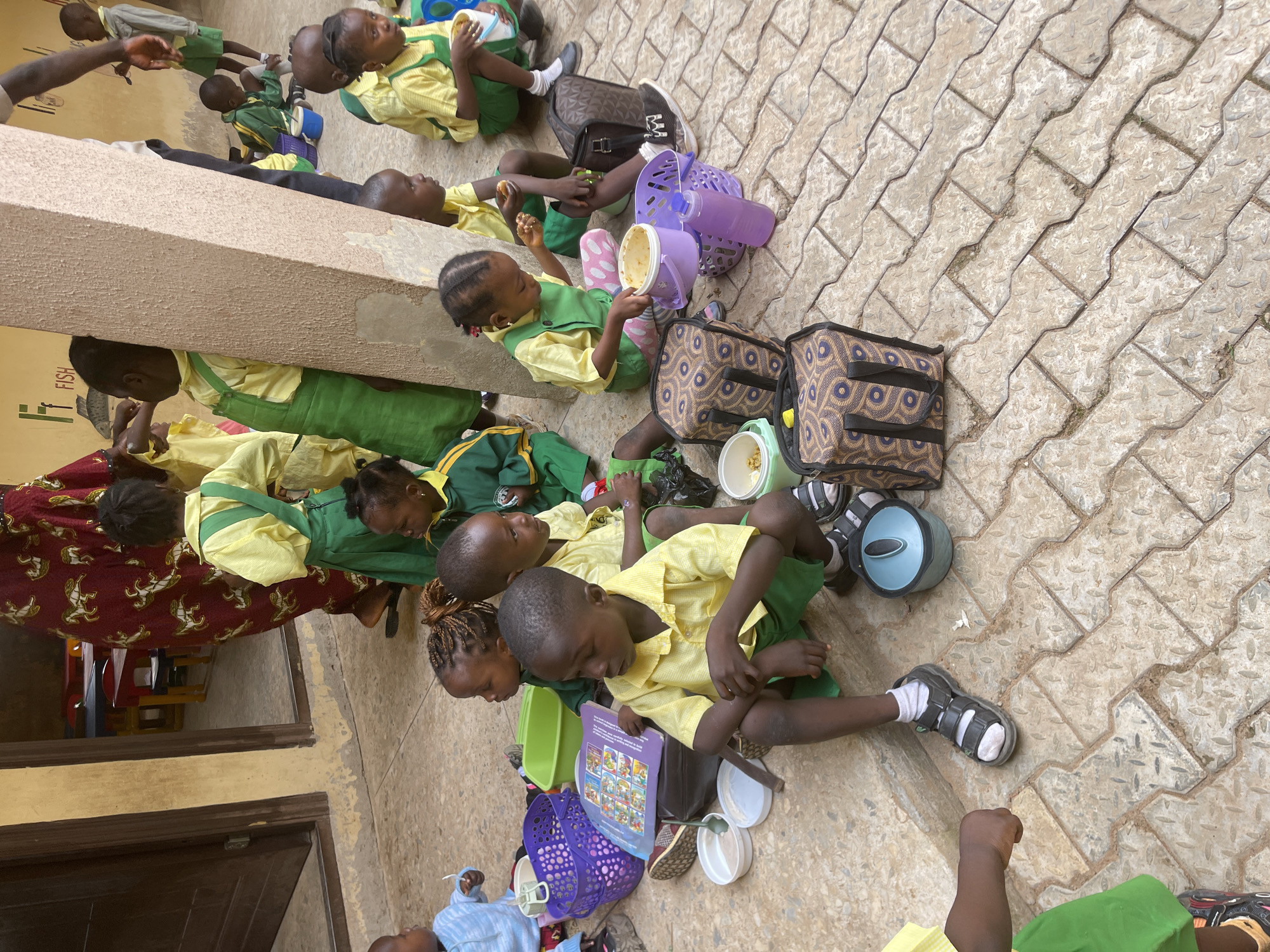 In Akwanga, Nigeria, a group of young schoolchildren, appearing to be of primary school age, are gathered outdoors on a paved area. They are dressed in school uniforms consisting of yellow shirts and green overalls or skirts. Many of them are seated on the ground, some with lunchboxes and containers, suggesting they are either eating or preparing to eat.
In the foreground, a young boy in a yellow shirt and green shorts is sitting cross-legged, looking down at a purple book. To his left, a purple basket and several small white bowls are visible. Further to the right, another child is reaching into a purple container. Several patterned bags are scattered around the children.
In the background, other children in similar uniforms are also seated. One child is holding a yellow cup, and another appears to be eating. A partially visible adult figure in a dark patterned garment is standing near a doorway. The environment includes a textured wall, a wooden door, and hints of a building interior with tables and chairs. The overall scene suggests a school setting, possibly during a lunch break or outdoor activity.