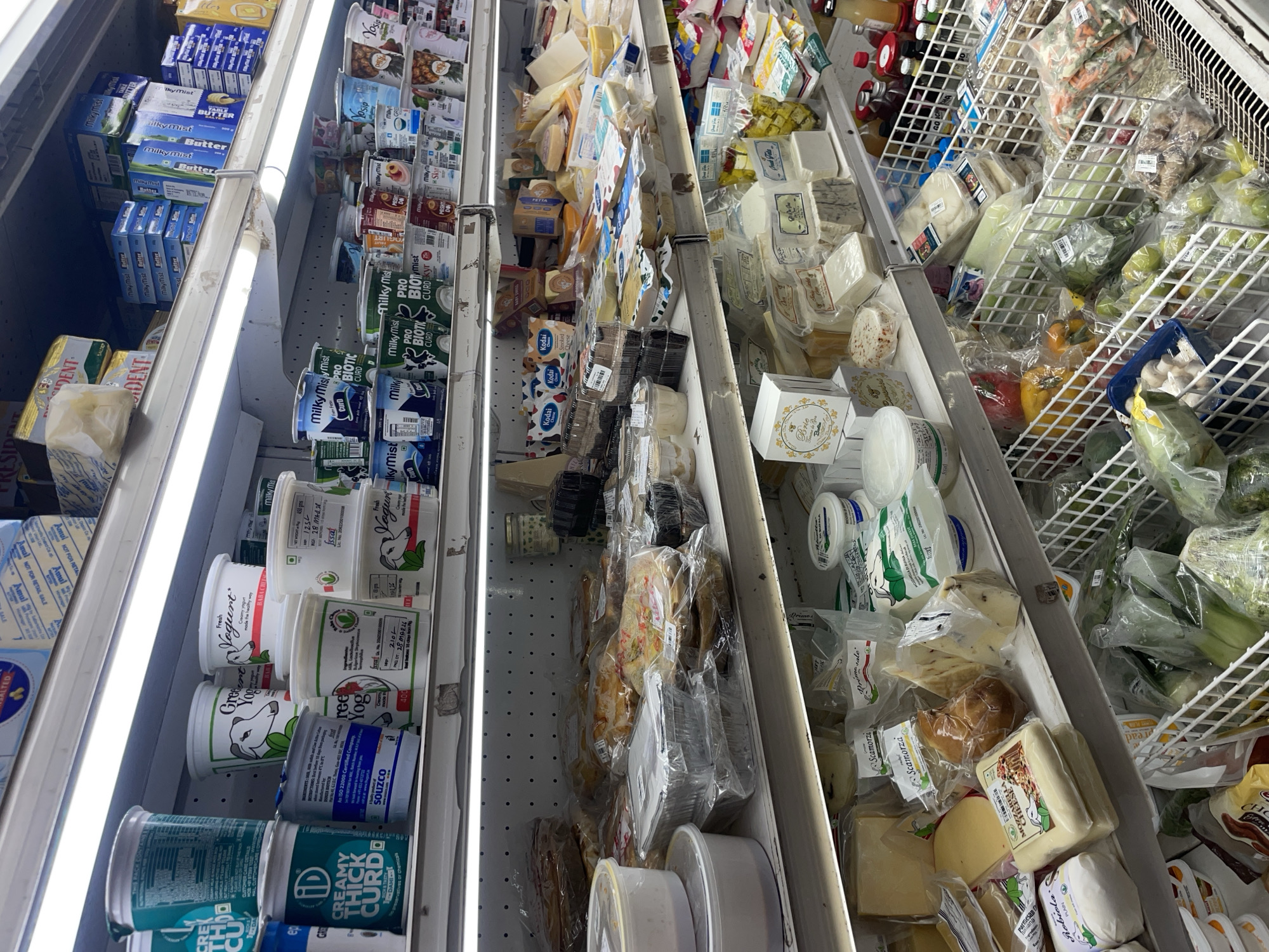 The image shows a refrigerated section in a store, likely in Oxel, India given the presence of brands such as Amul, Milky Mist, and Souzco. The shelves are stocked with a variety of dairy products including butter, yogurt, curd, and cheese. Vegetables and prepackaged salads are visible in the far right section of the refrigerator. The shelves are brightly lit with LED lighting.