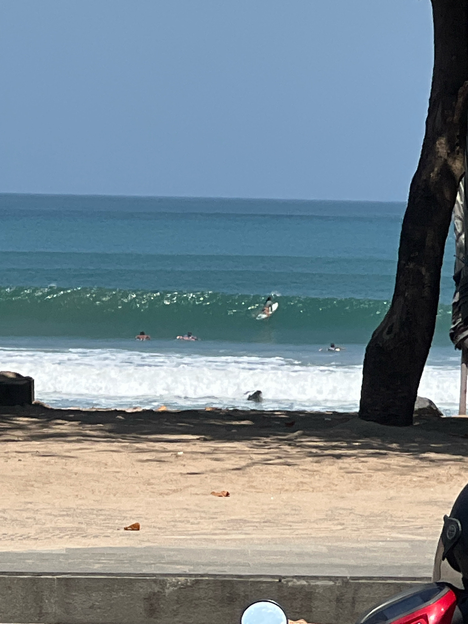 In Kuta, Indonesia, on a sunny day with clear skies, the ocean provides a backdrop of turquoise water and breaking waves. Several surfers are visible in the water, some riding waves and others waiting for them. The foreground features a sandy beach with shadows cast by trees, and a paved area in front of a wall where a motorcycle is parked. The scene captures a typical day at the beach in Kuta.