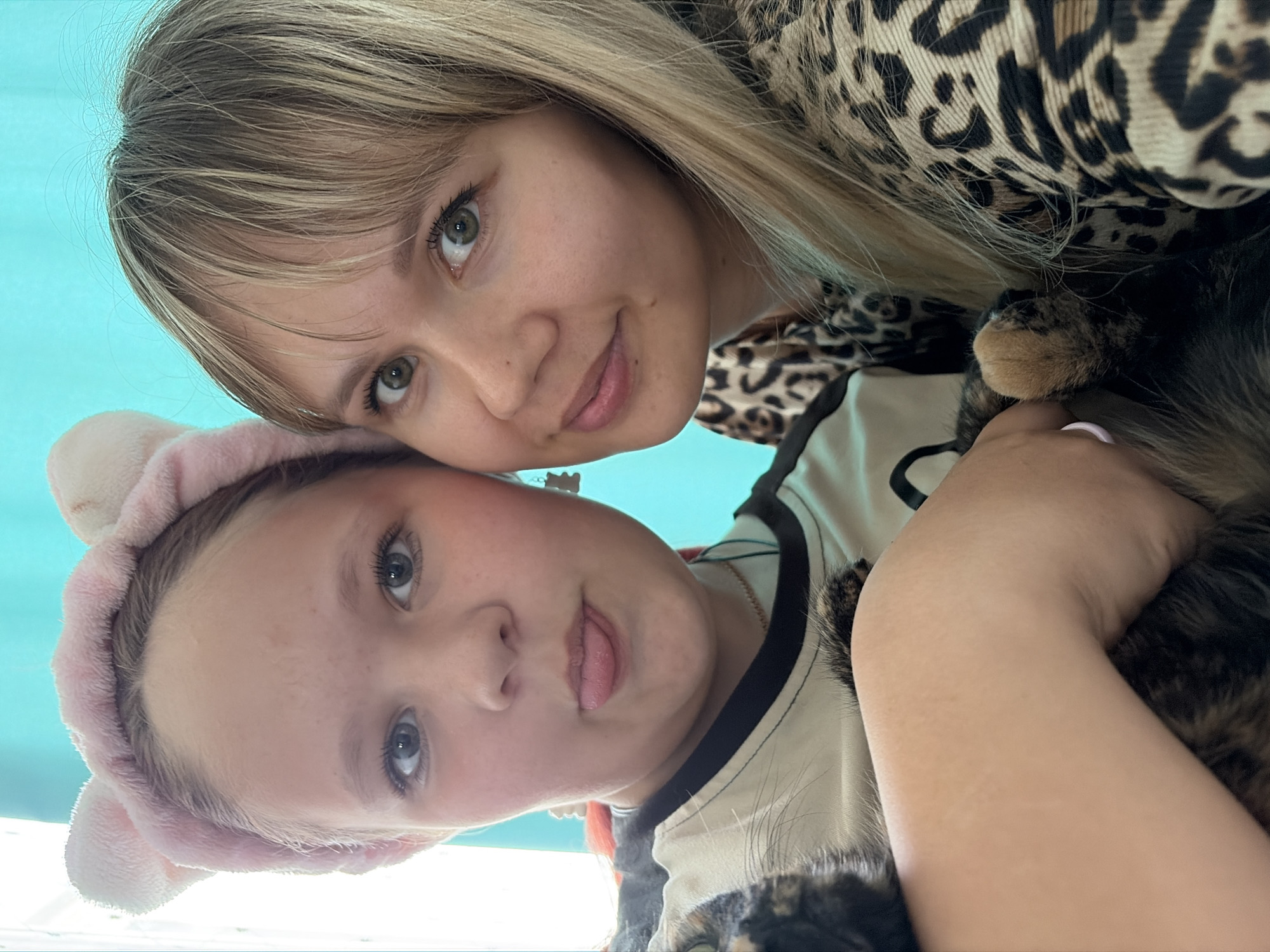 Two females, one older and one younger, are posed in a close-up selfie. The older female, wearing a leopard print garment, is positioned above the younger female. The younger female wears a pink headband with ears and a t-shirt with black trim and a graphic of a cat. Both are looking at the camera. Part of a cat is seen in the arms of the younger female. The background is a light blue color. There is no specific evidence in the image to confirm the location as Chaikovsky, Russia. The photograph seems to have been taken indoors.